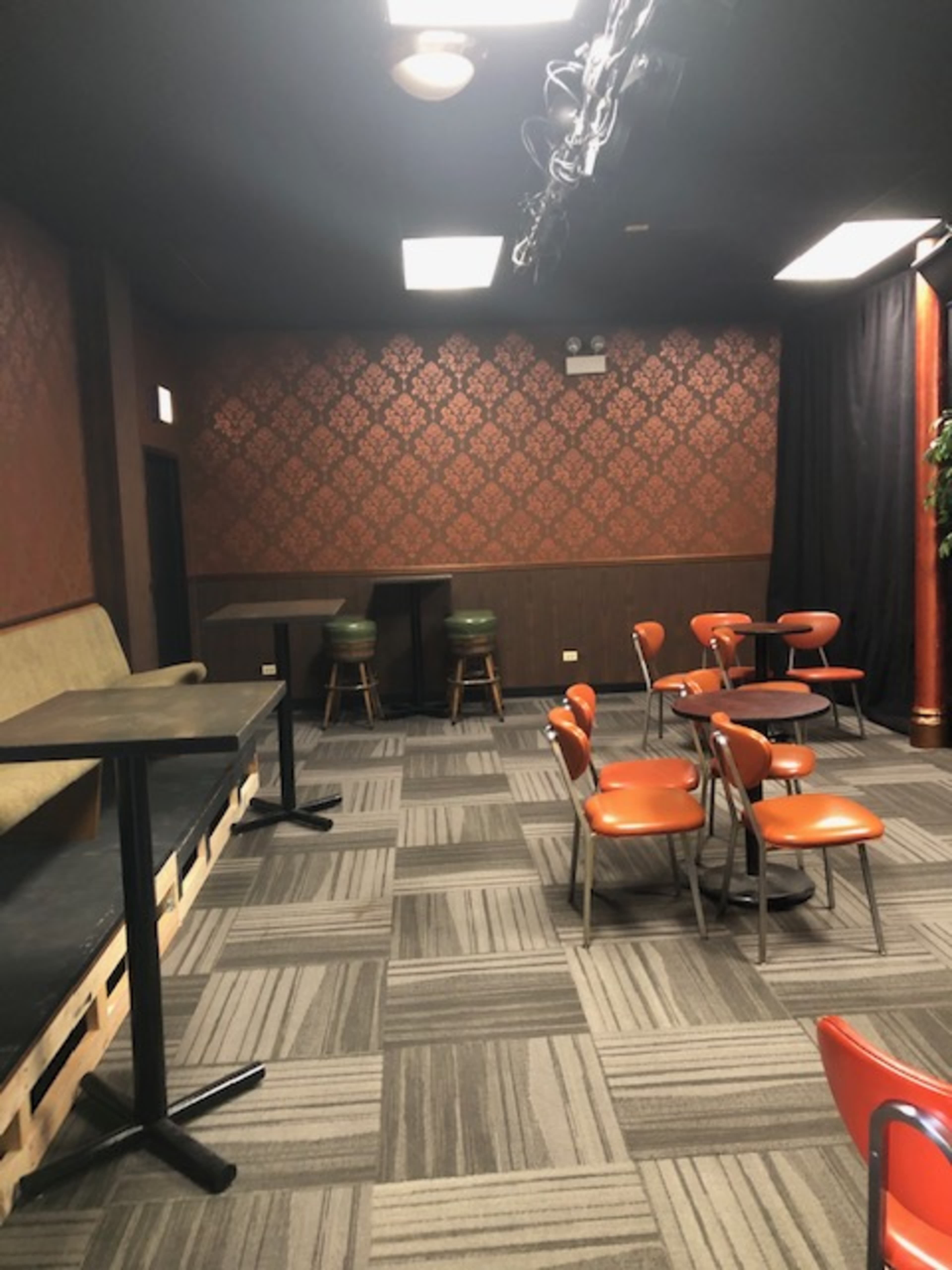 A sparsely furnished room with orange chairs arranged around small tables, a couch along one wall, and a bar area with stools in the background.