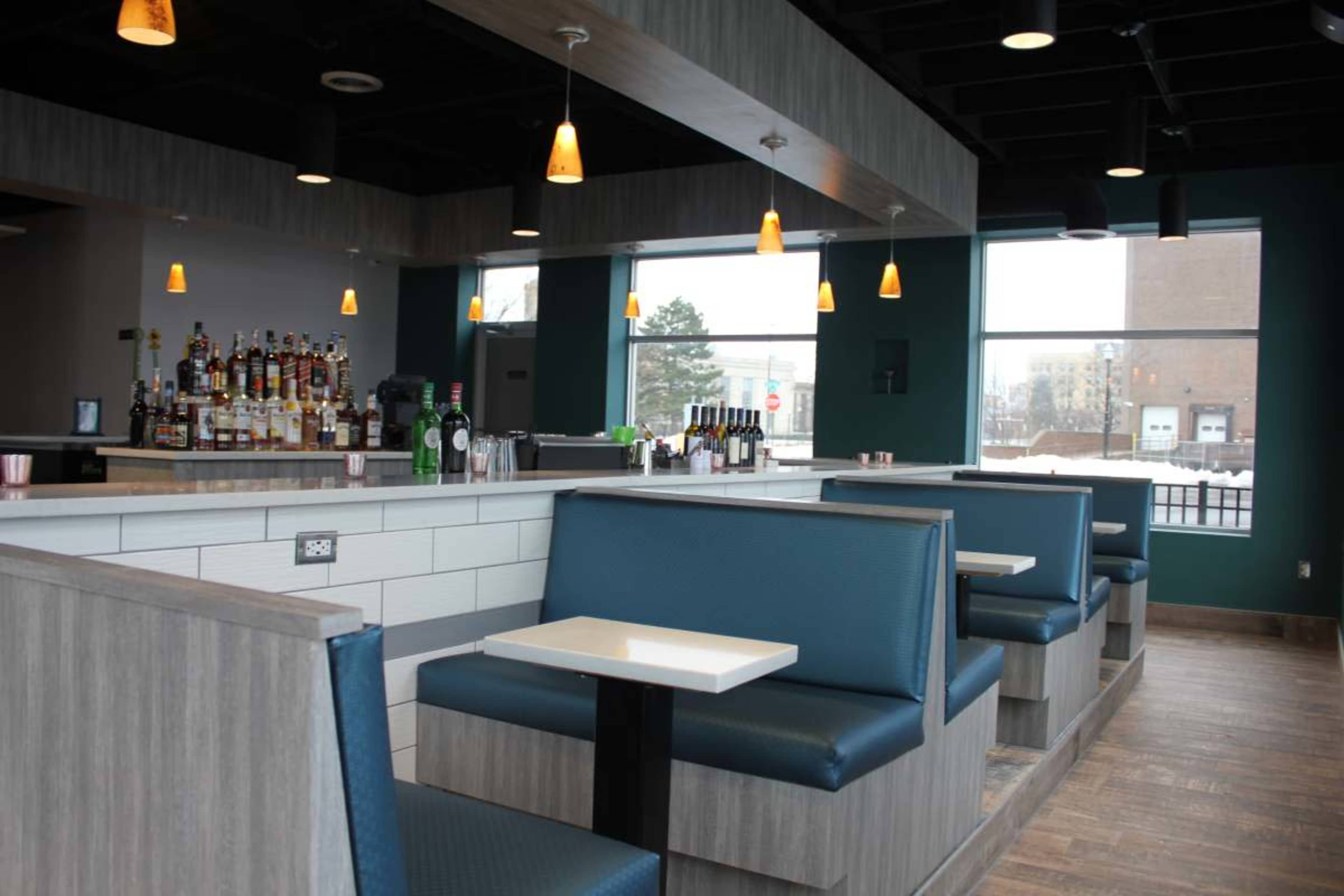 The image shows the interior of a restaurant featuring booths with blue seating, a bar area with various bottles, and large windows allowing natural light.