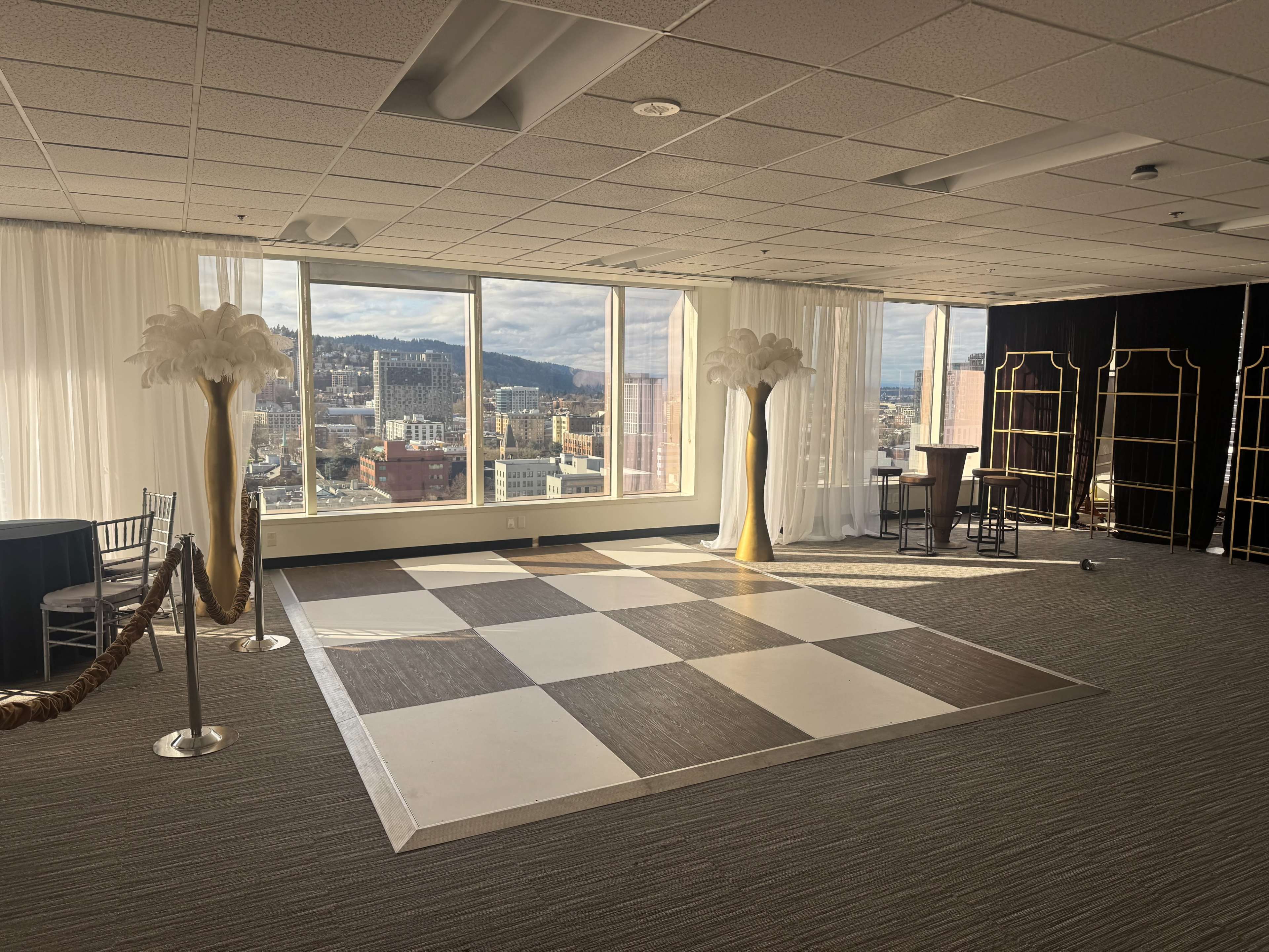 The image shows a spacious event room featuring a checkered dance floor, tall decorative vases with feathers, and large windows with a cityscape view in the background.