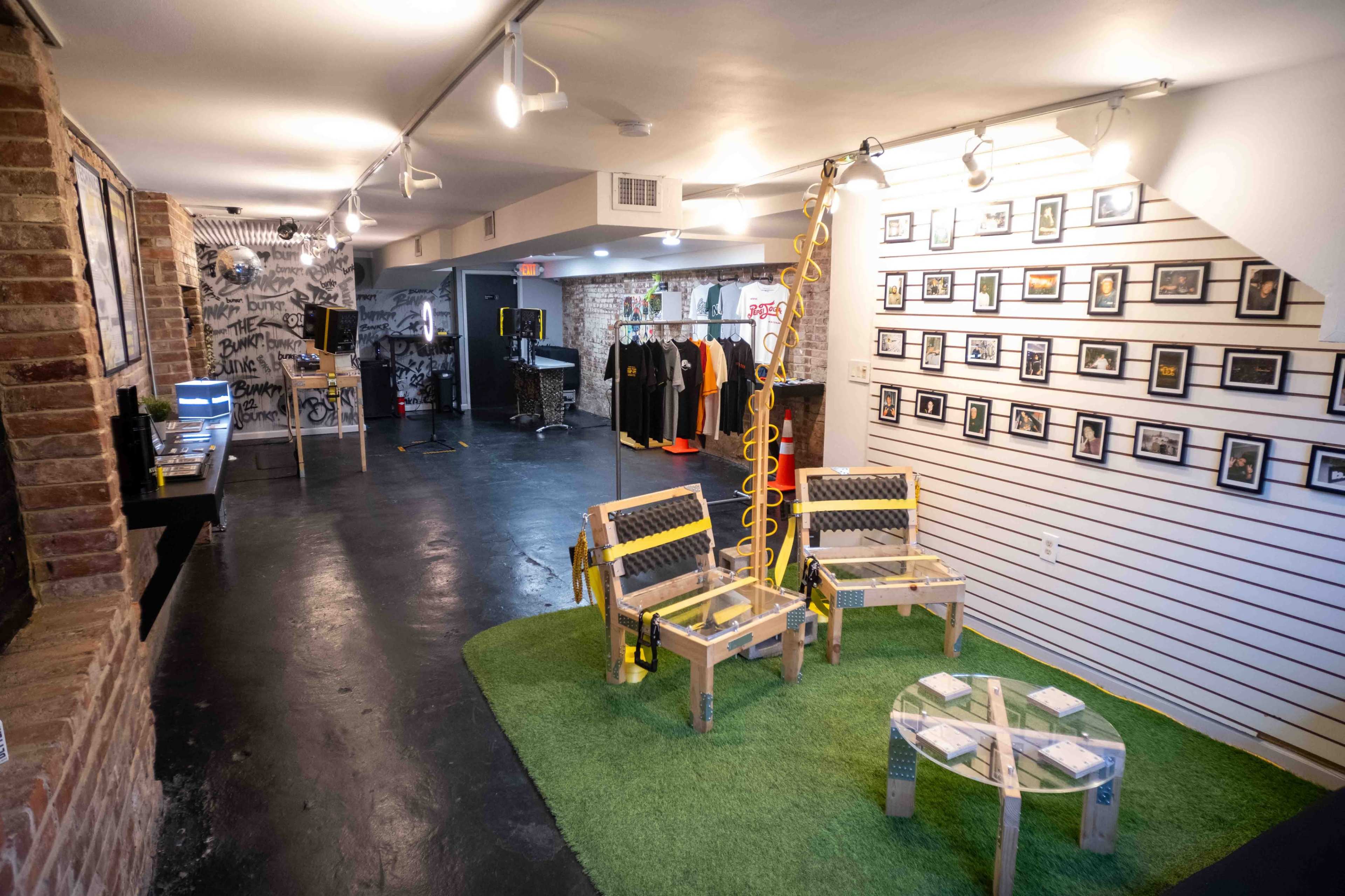 An indoor space with exposed brick walls, featuring a seating area with two wooden chairs on green grass-like flooring, a display of photographs on one wall, and clothing hanging in the background.