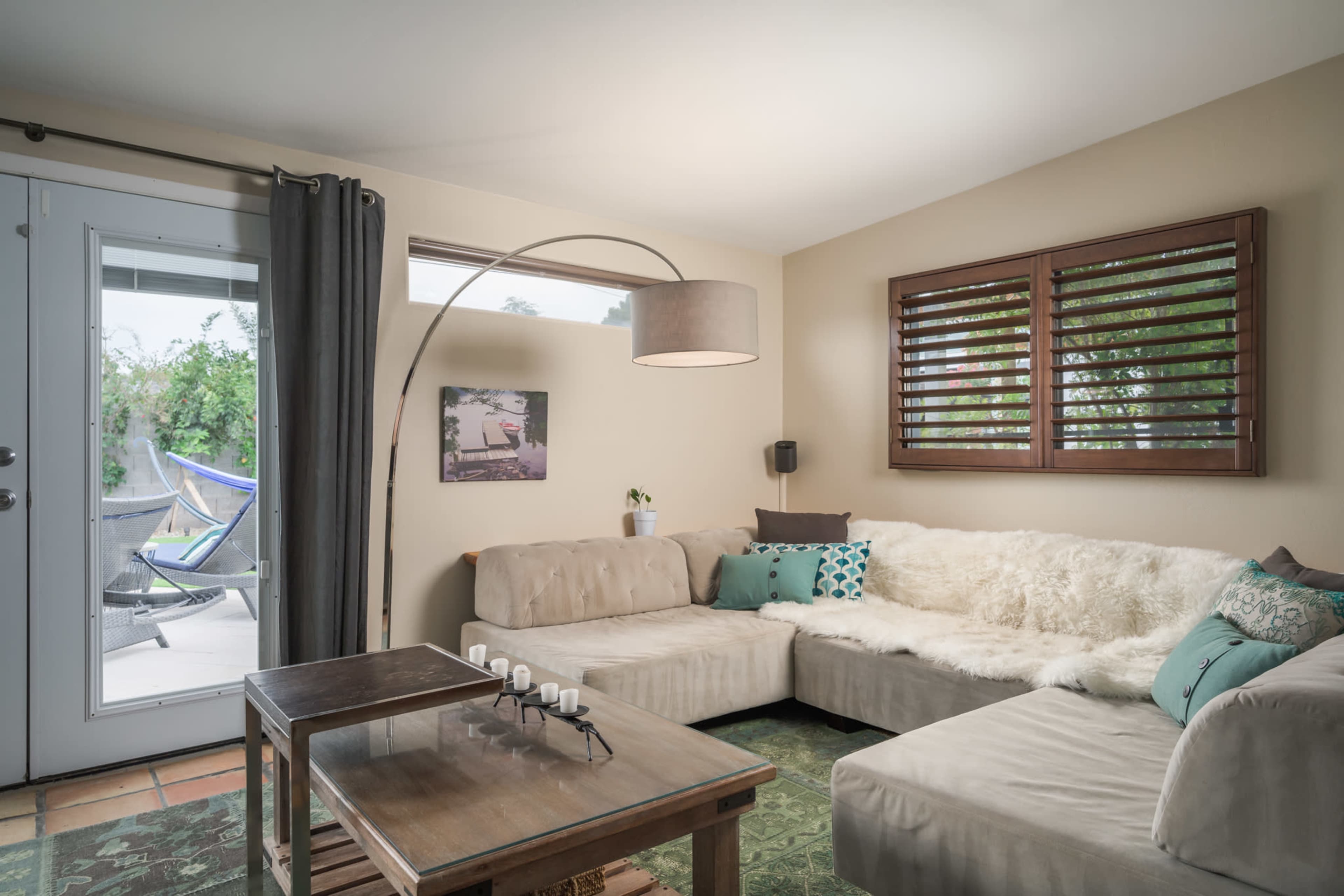 A cozy living room features a sectional sofa, a wooden coffee table, and a large window with shutters, opening to an outdoor patio area.