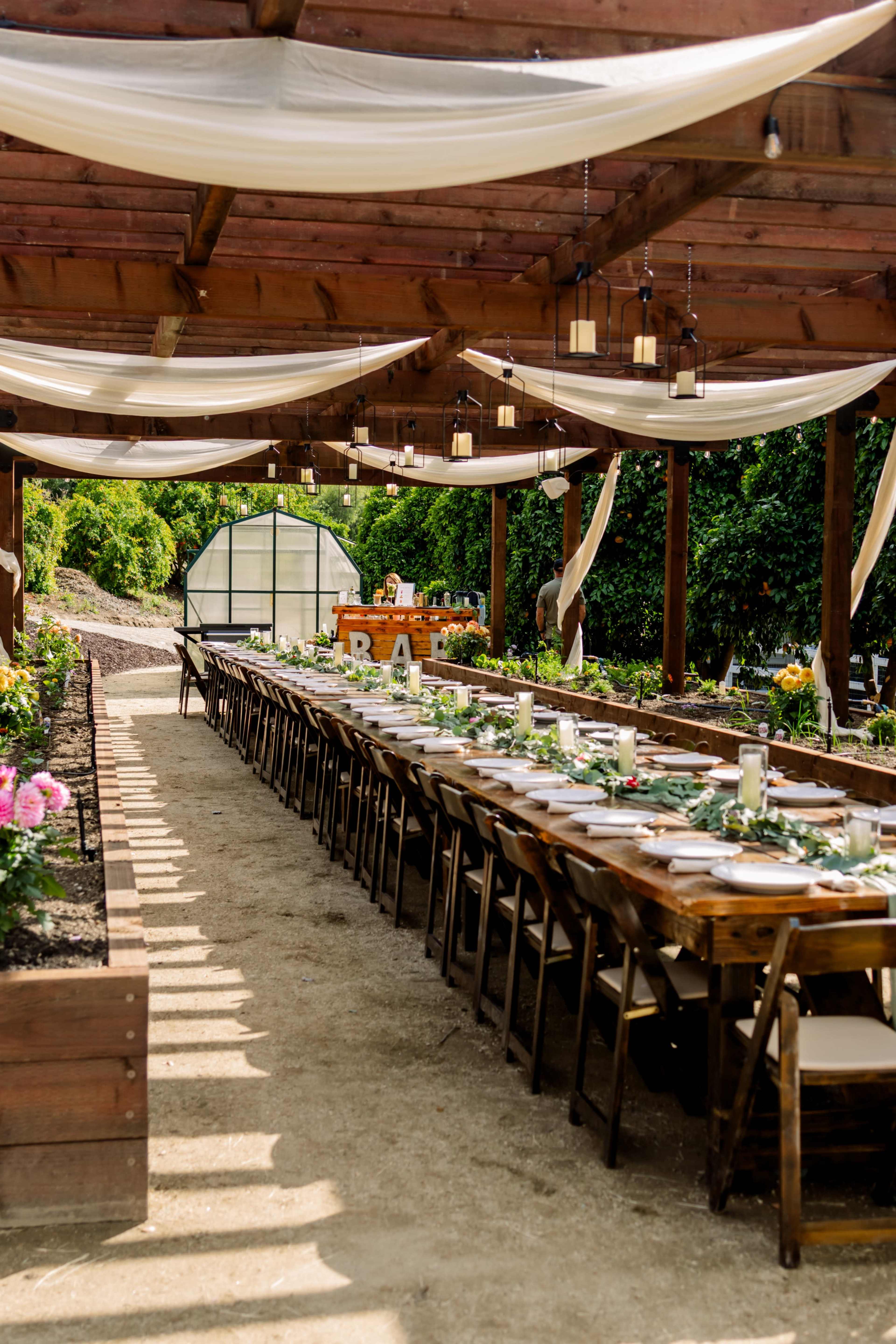 A long wooden table is set for an outdoor wedding reception under a wooden pergola, with white drapes and lanterns overhead.