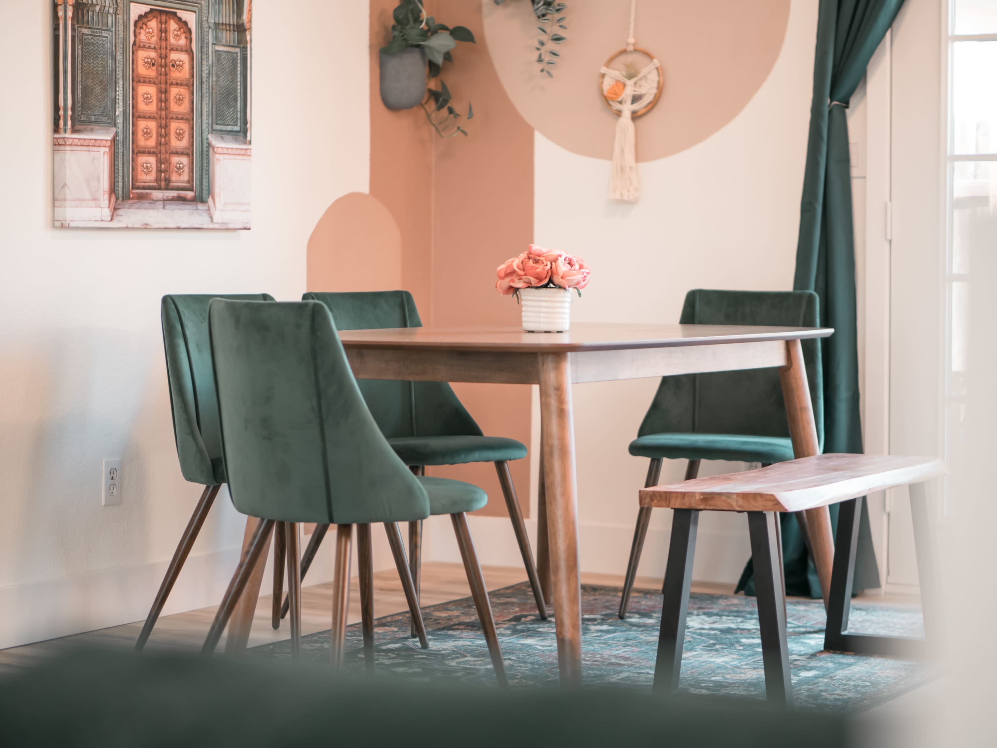 A wooden dining table is surrounded by four green velvet chairs, with a bouquet of pink flowers in a white vase on the table, set against a backdrop of light-colored walls and decorative accents.