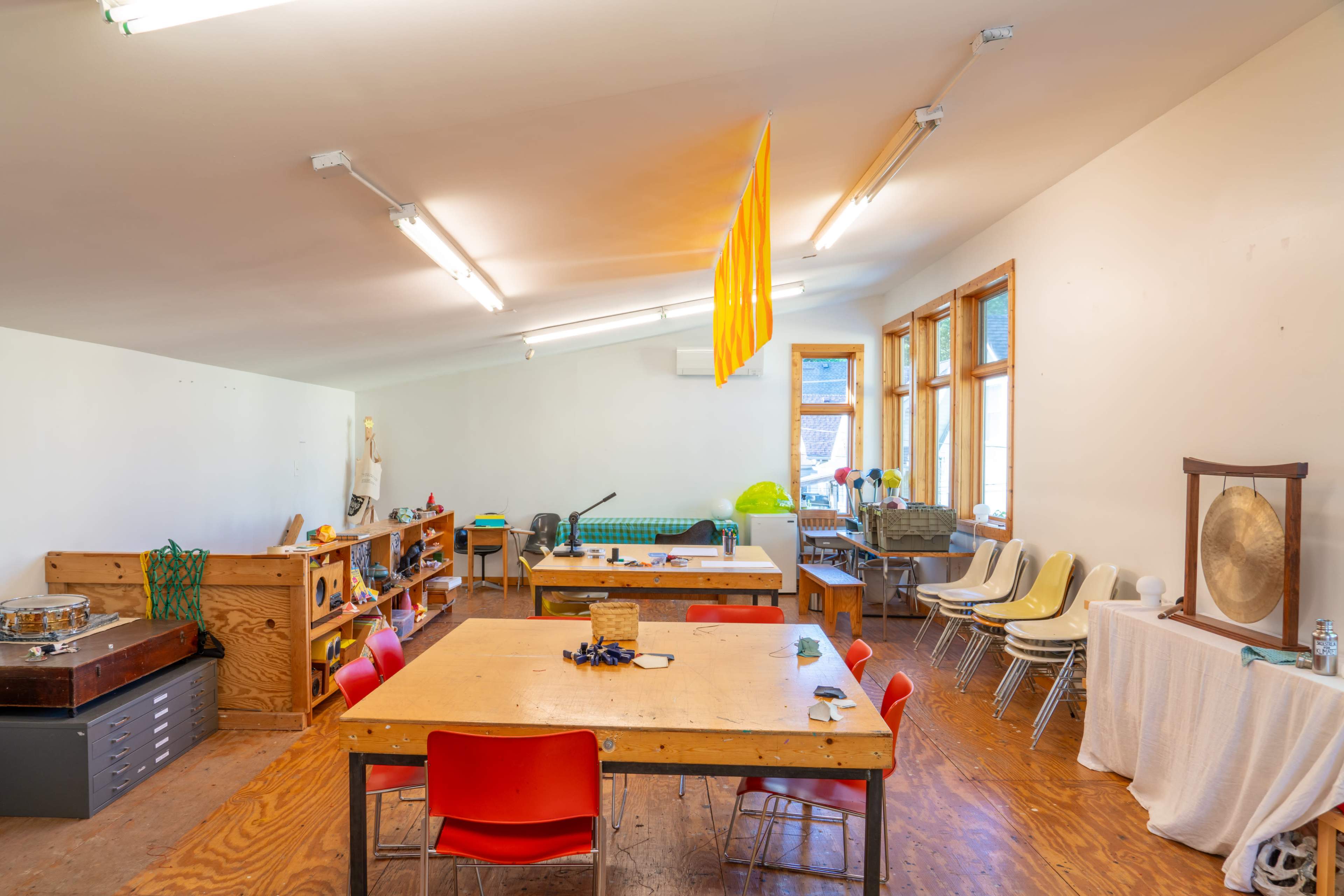 A spacious art studio with wooden tables, colored chairs, large windows, and various creative supplies arranged throughout the room.
