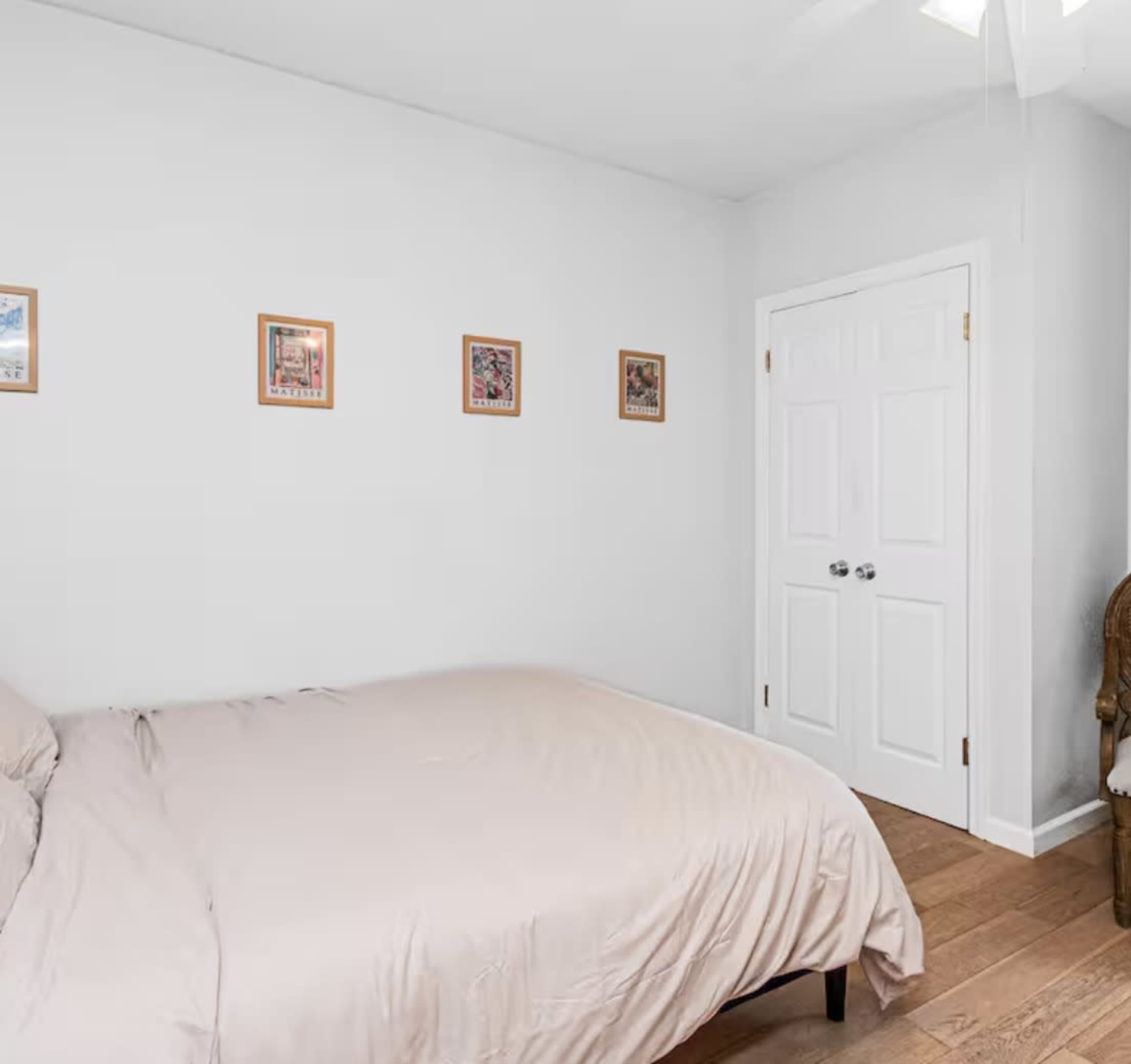 A simple bedroom features a bed with a light-colored duvet, framed pictures on the wall, and a door leading to another room.