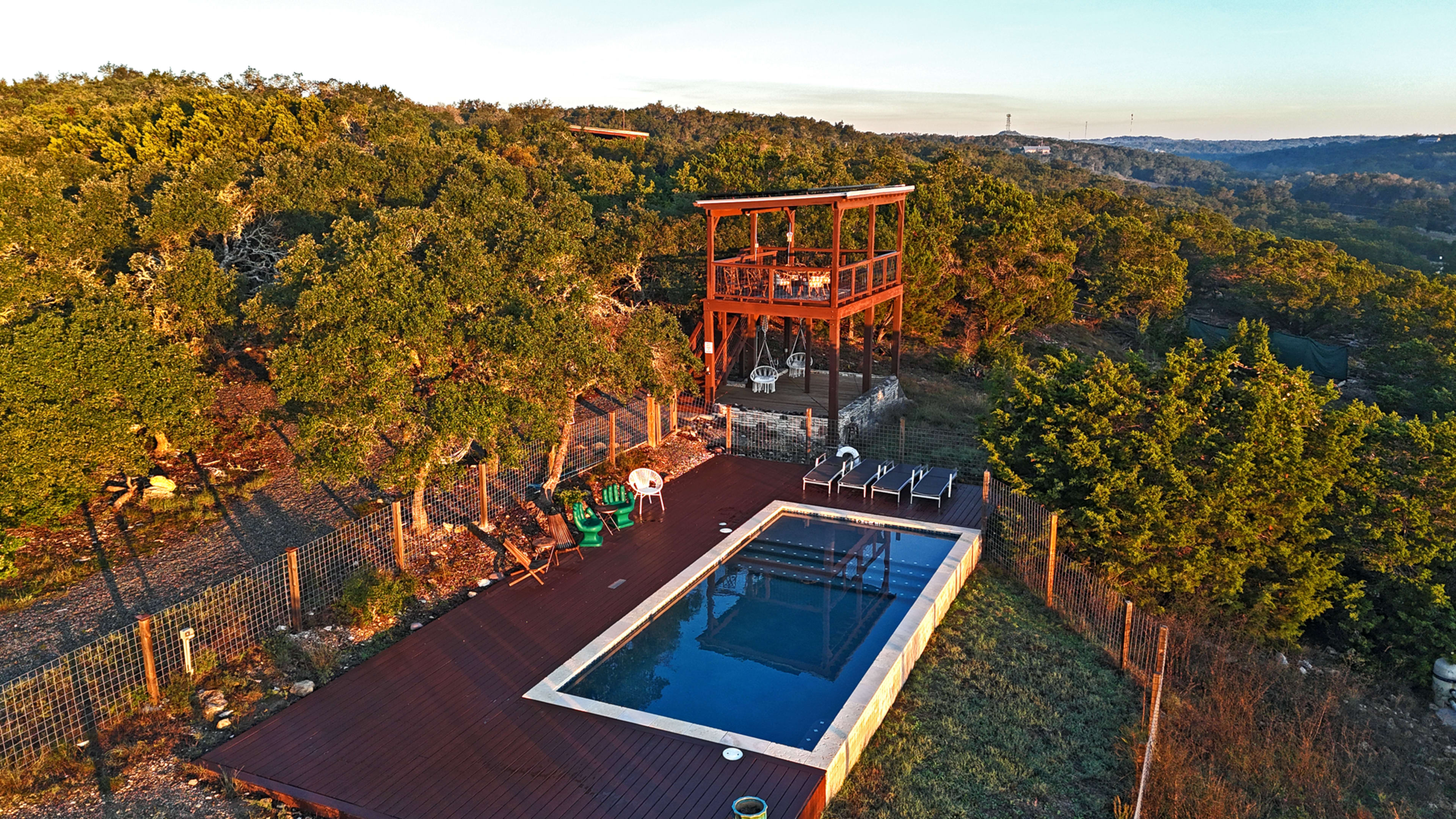 The image shows a wooden deck surrounding a rectangular swimming pool, located on a hillside amid trees, with a multi-level observation tower overlooking the area.