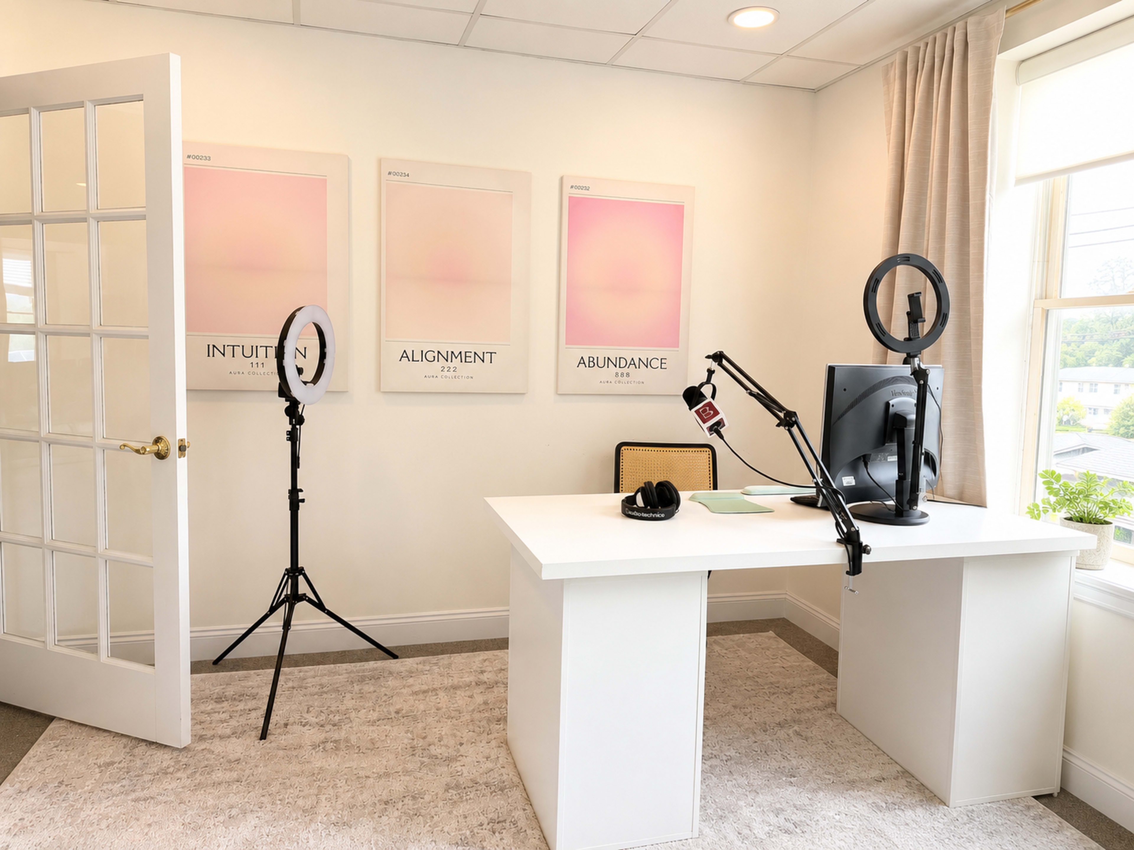The image shows a neatly organized office space featuring a white desk, a microphone on a stand, a ring light on a tripod, and three framed posters on the wall.