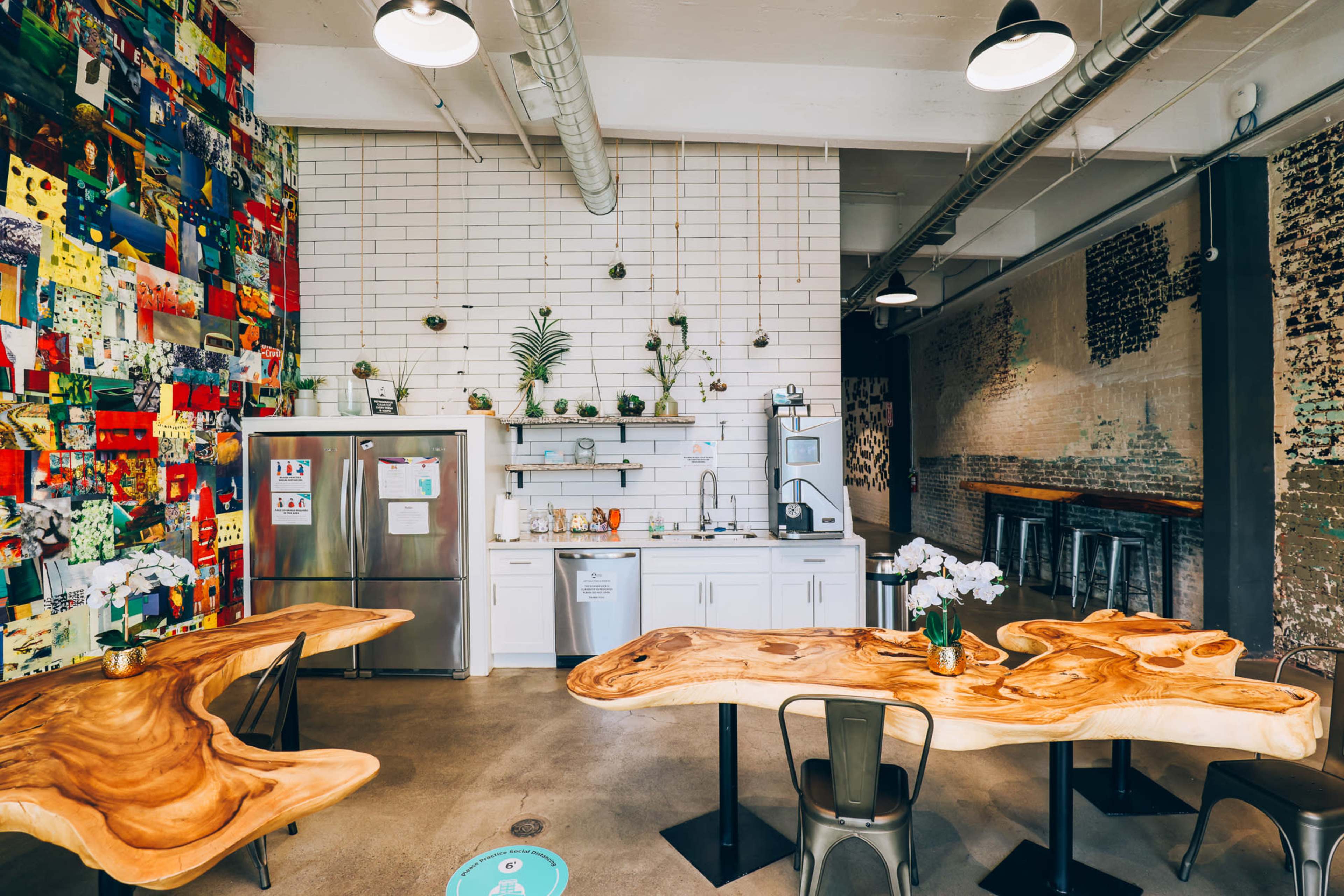 The image shows a modern kitchen area with wooden tables and a colorful wall featuring an array of decorative artworks.