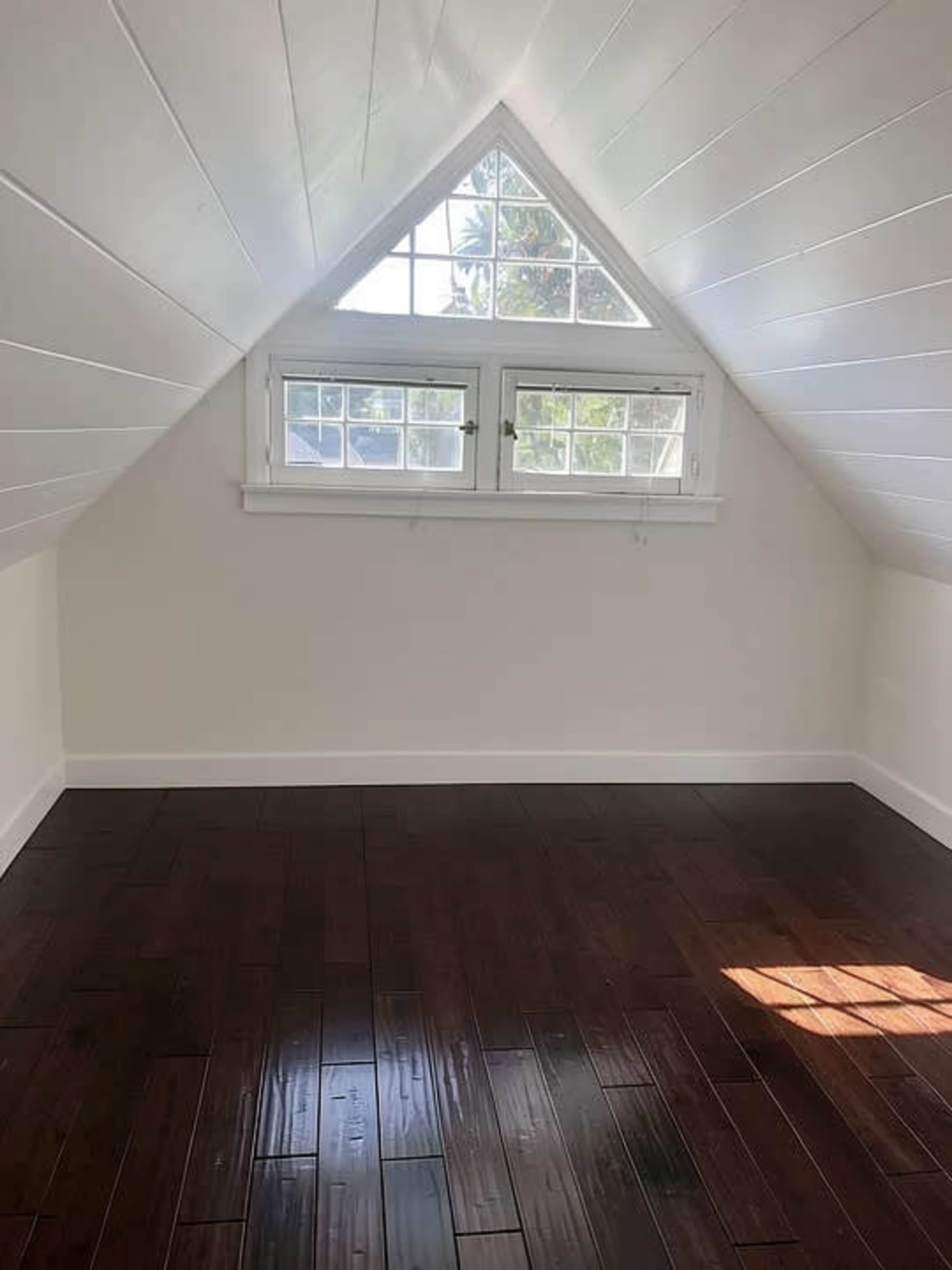The image shows a small, empty attic room with wooden flooring and a triangular window at the apex of the sloped ceiling.