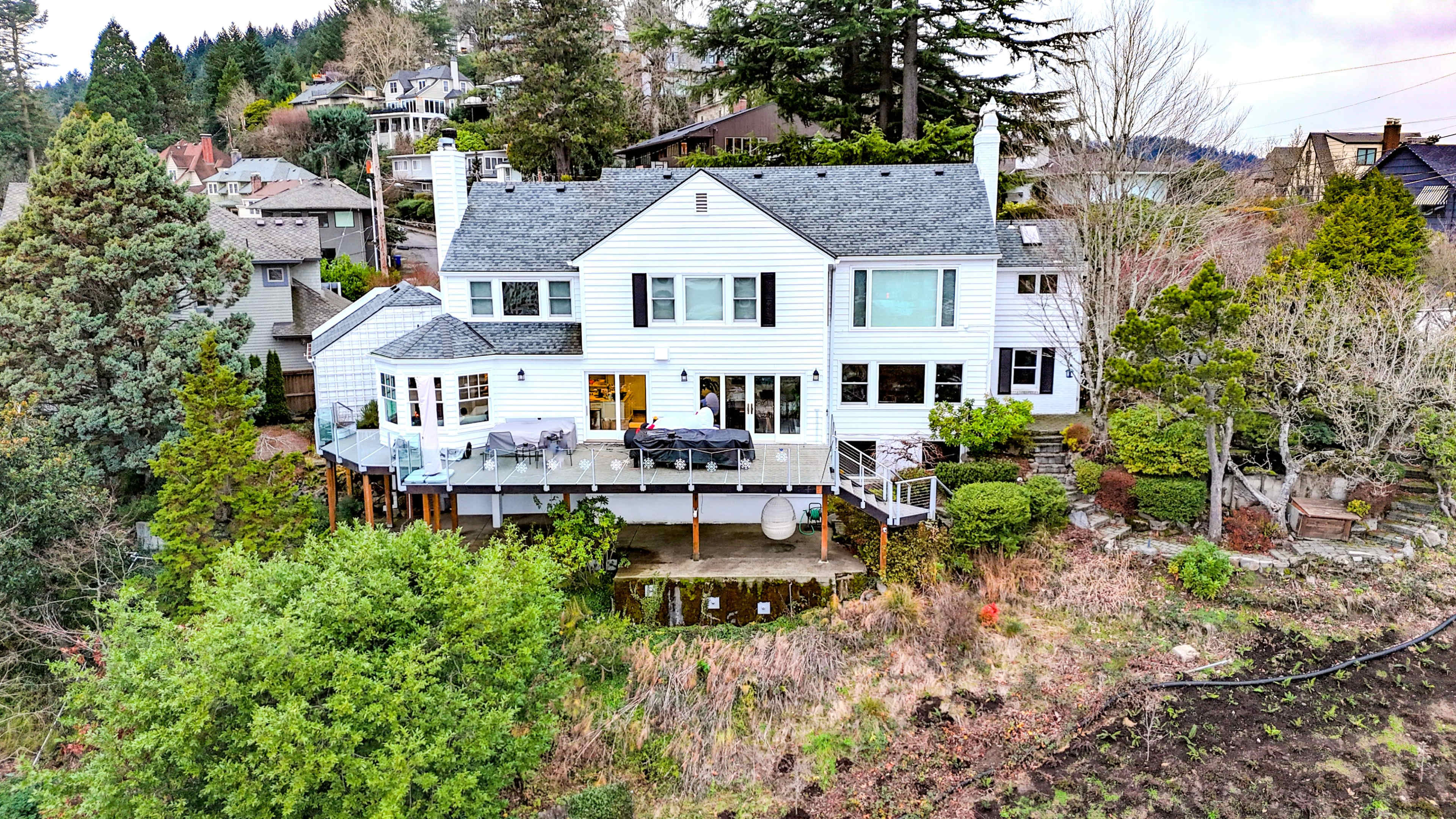 A two-story white house with a large wooden deck is situated on a hillside, surrounded by trees and shrubs.
