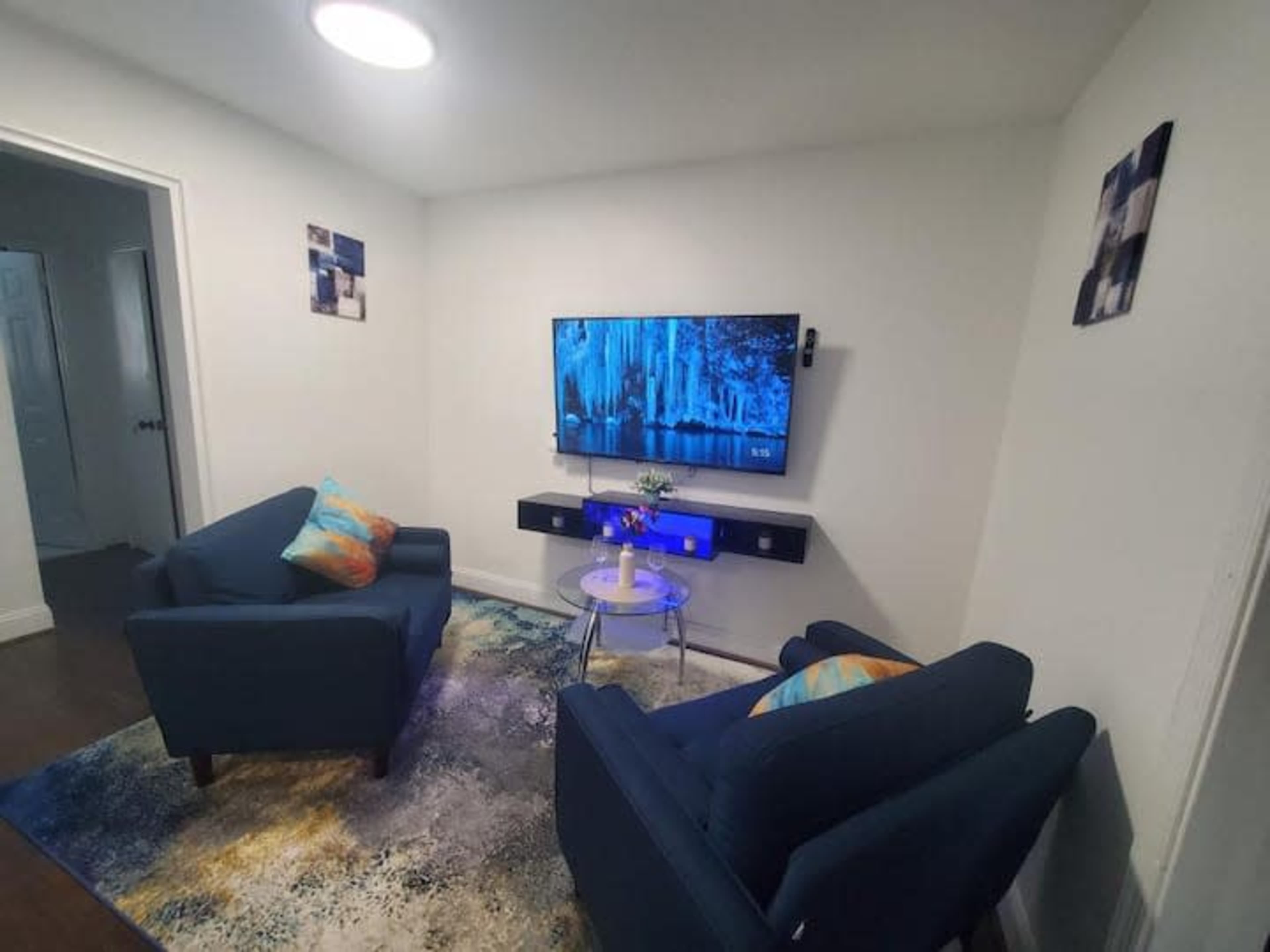 The room features two blue chairs facing a wall-mounted television, with a small table and a decorative rug on the floor.