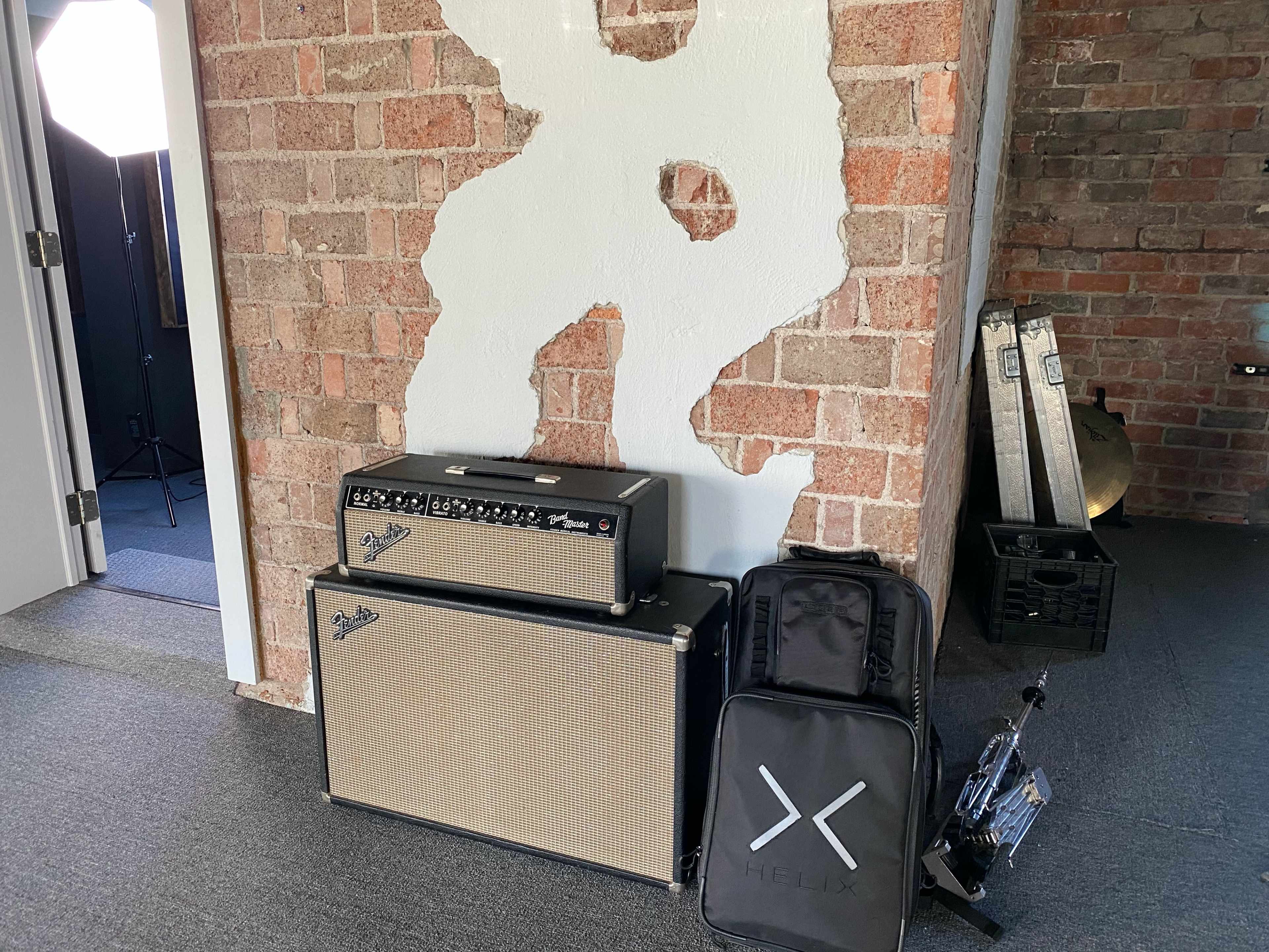 A guitar amplifier and a backpack sit against a brick wall in a room with a gray carpet.