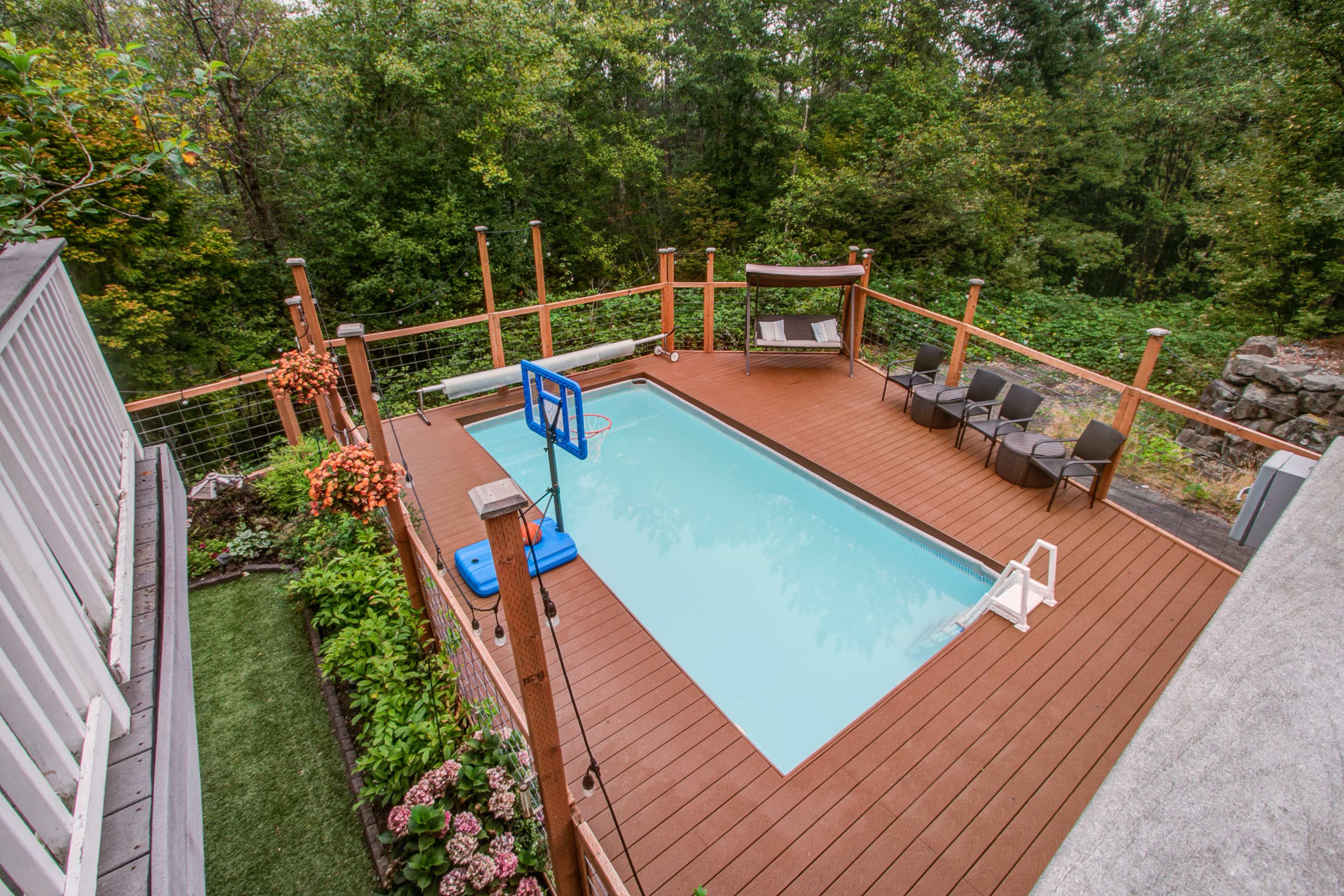 The image shows a rectangular swimming pool surrounded by a wooden deck, seating chairs, and lush greenery in the background.