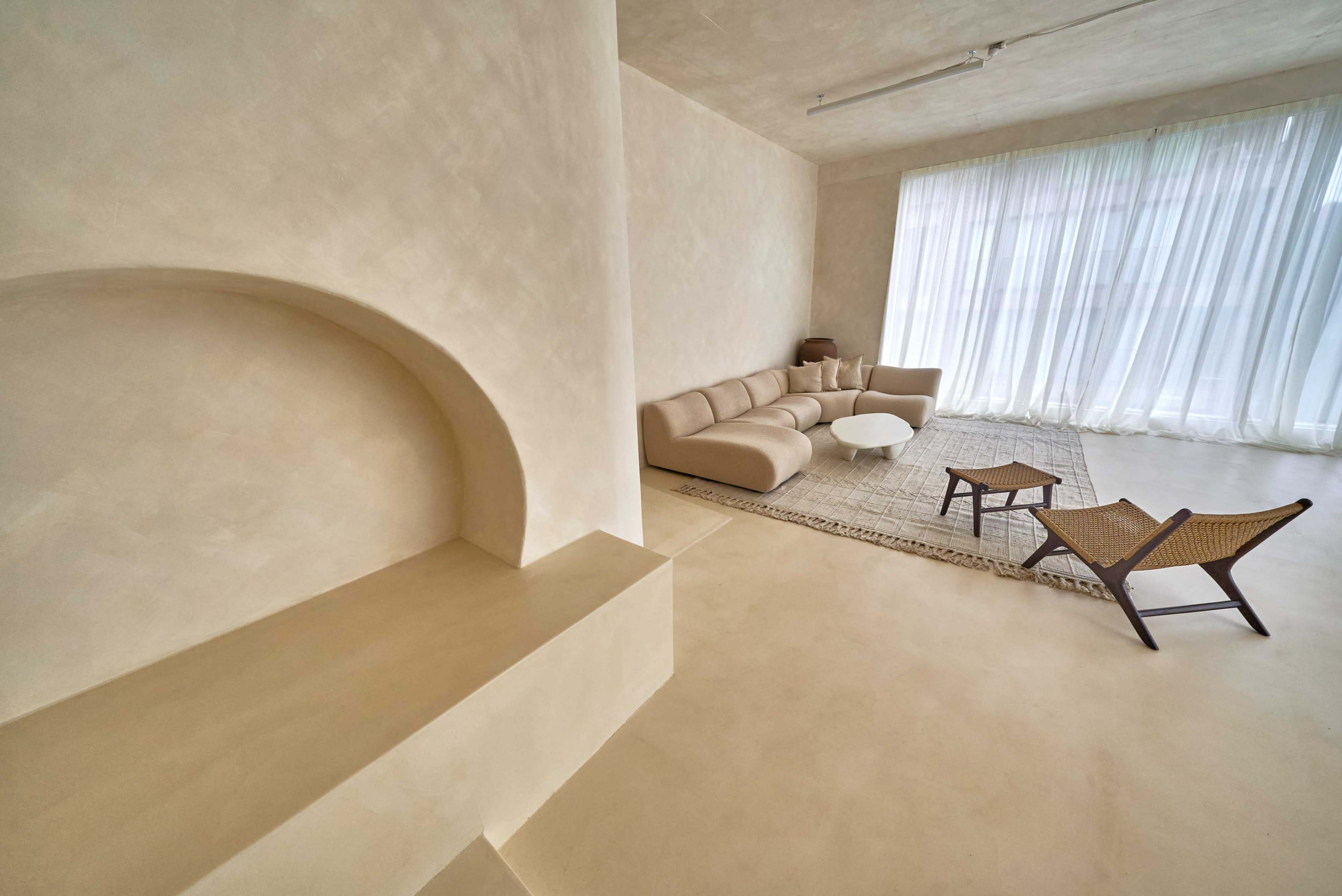 The image shows a minimalist living room with a beige sofa, a round coffee table, and a woven chair, all set against light-colored, textured walls and large windows covered with sheer curtains.