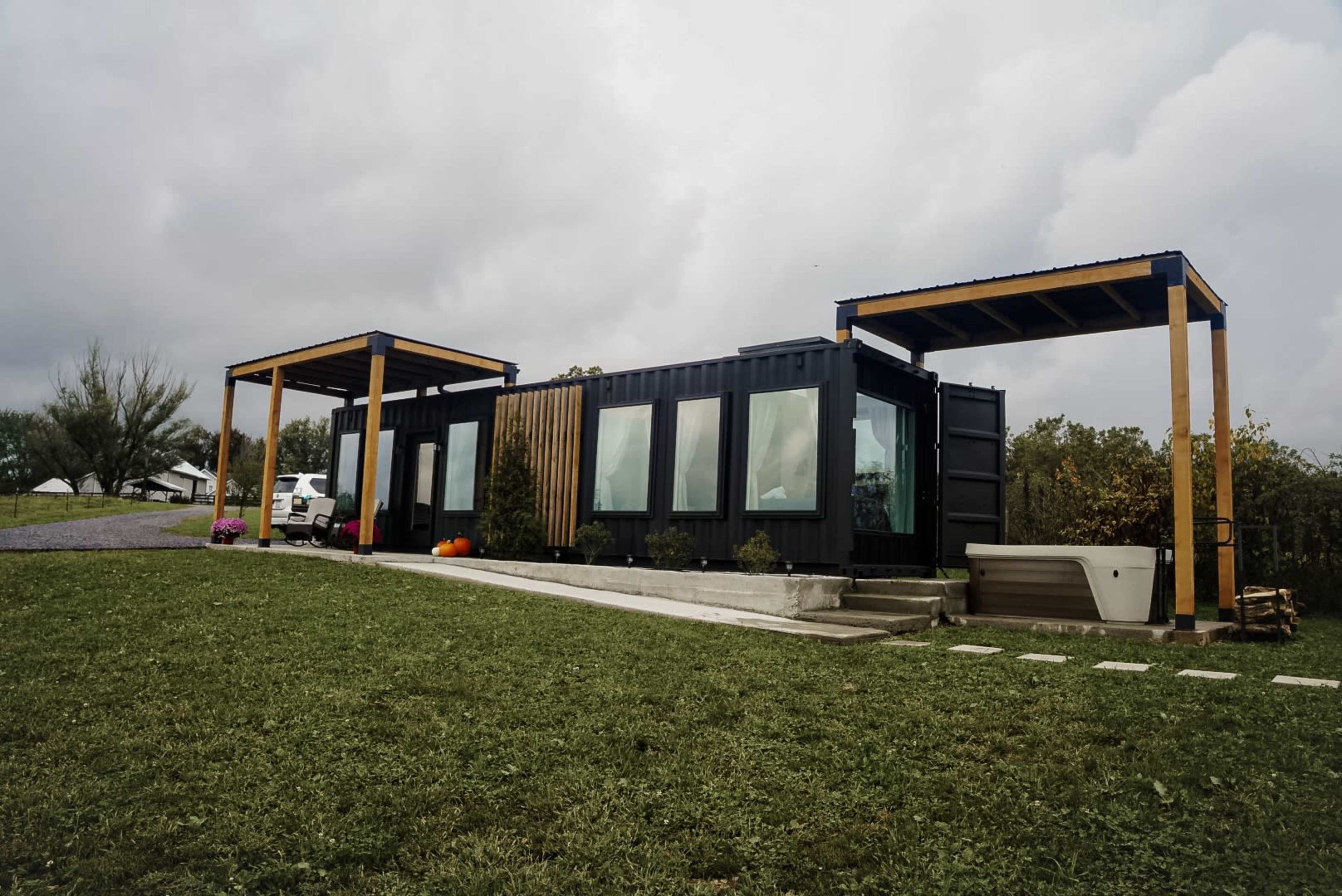 A modern container home with large windows and wooden overhangs sits on a grassy plot under a cloudy sky.