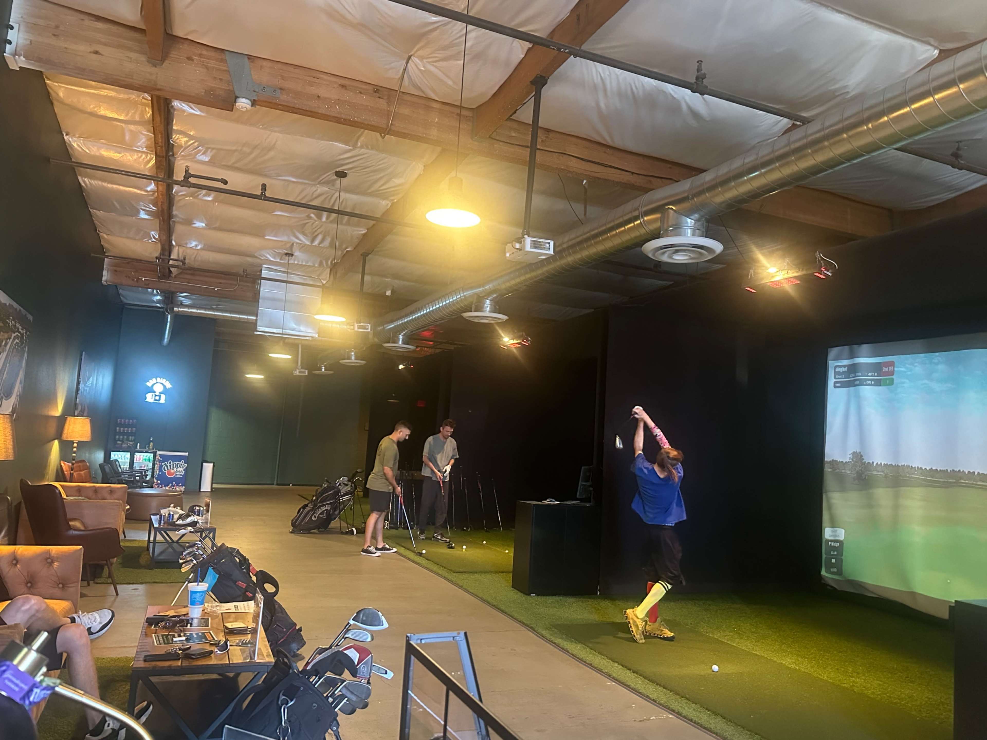 The image shows a golf simulation facility where several players are practicing their swings in front of a large screen displaying a golf course.