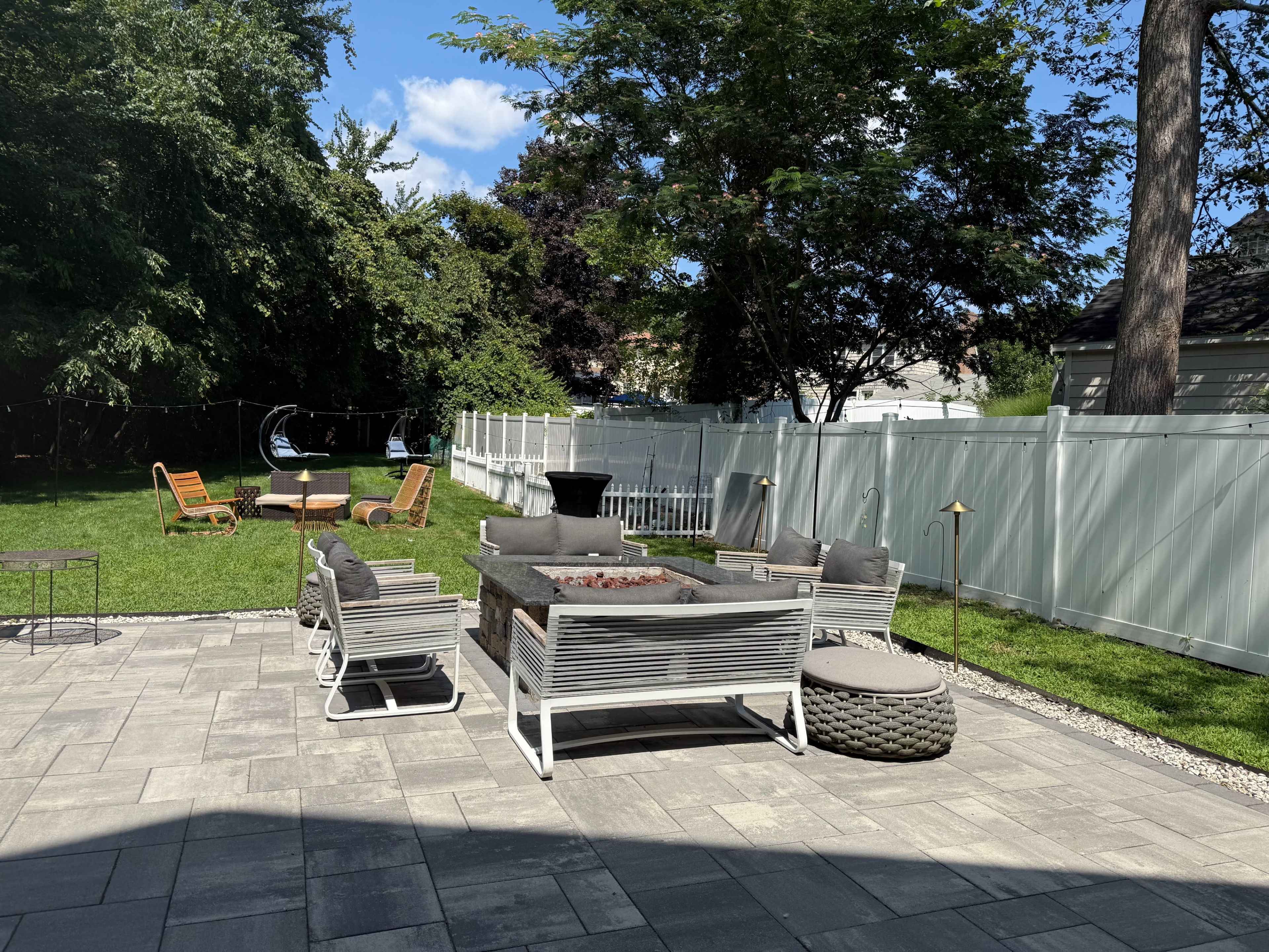 The image shows a backyard patio area with stone pavers, a fire pit surrounded by seating, and grassy spaces bordered by trees and a white fence.