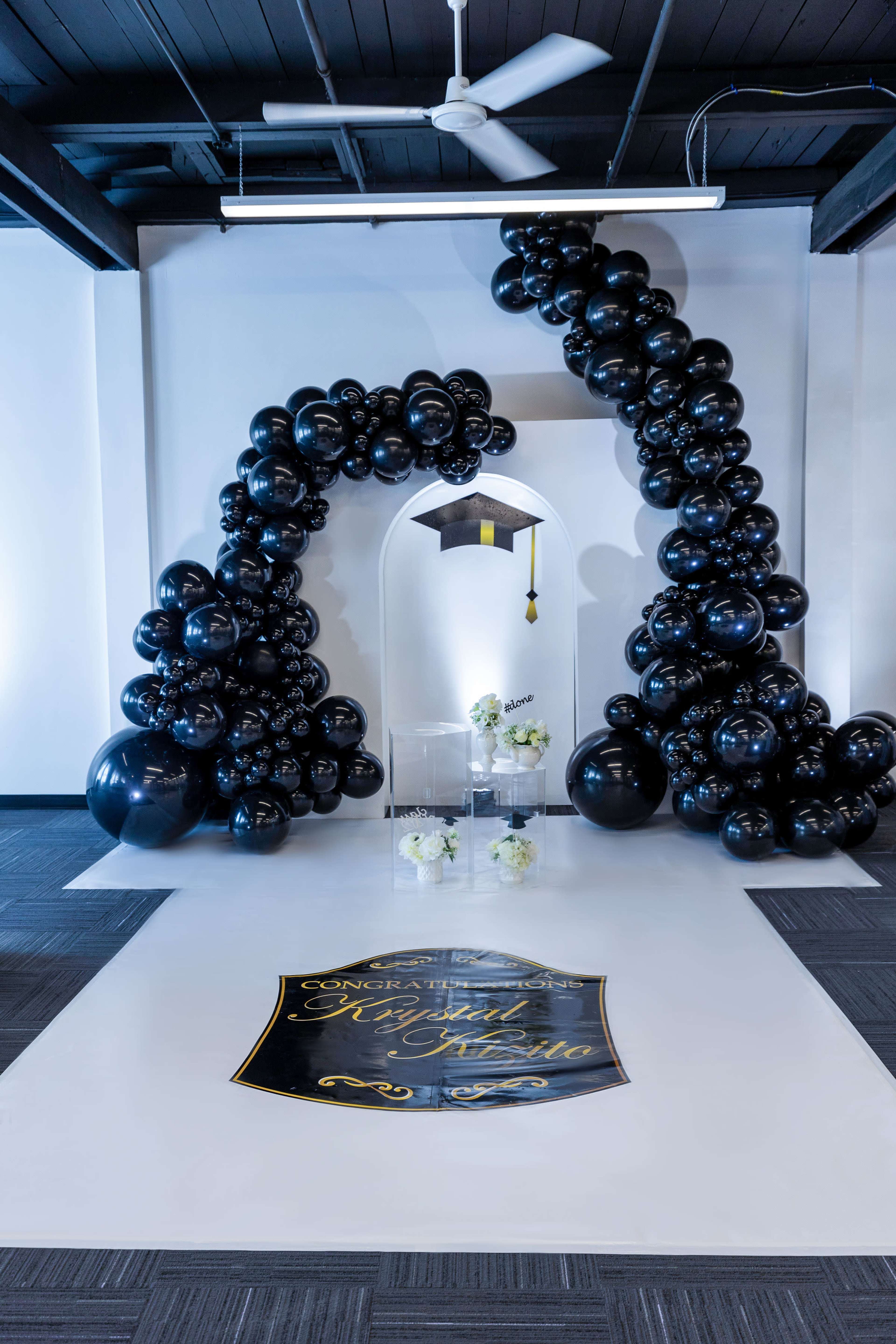 A decorative arch made of black balloons with a graduation cap at the top, set against a white wall, with a sign reading "Congratulations" on the floor.