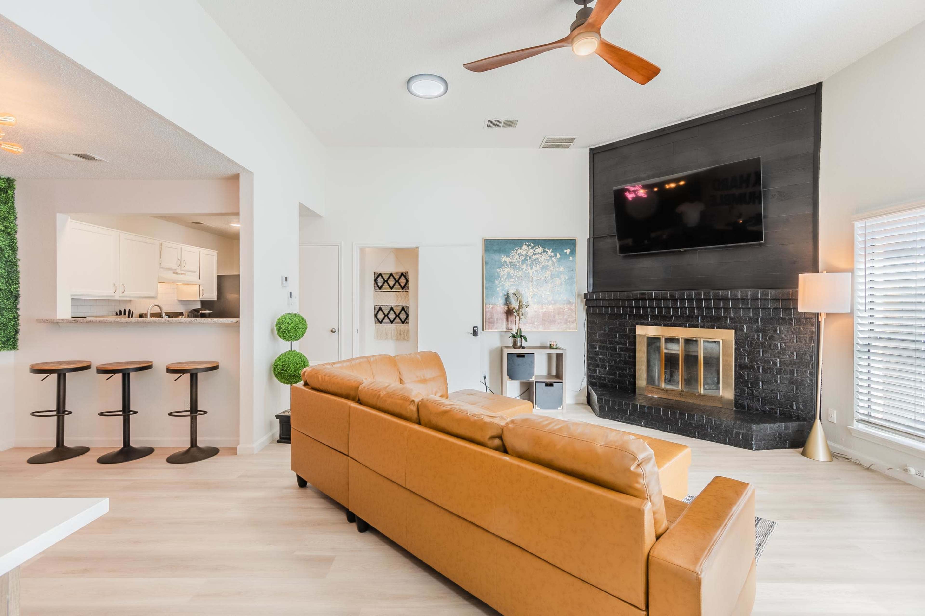 The image shows a modern living room featuring a brown leather sofa, a black fireplace, and a flat-screen TV mounted on the wall, with a kitchen area visible in the background.