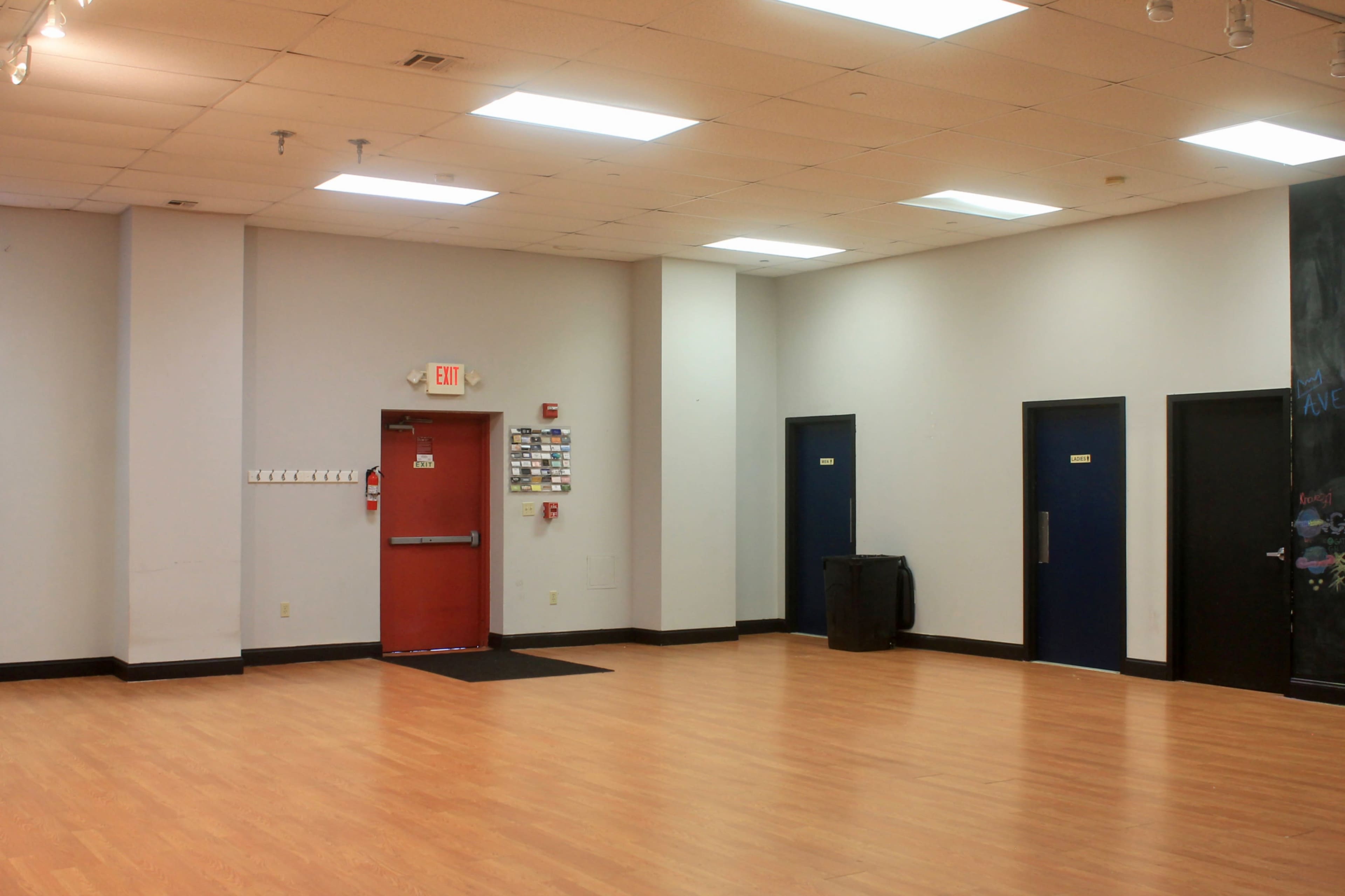 A spacious room with wooden flooring, featuring a red exit door and two blue doors on the right wall.