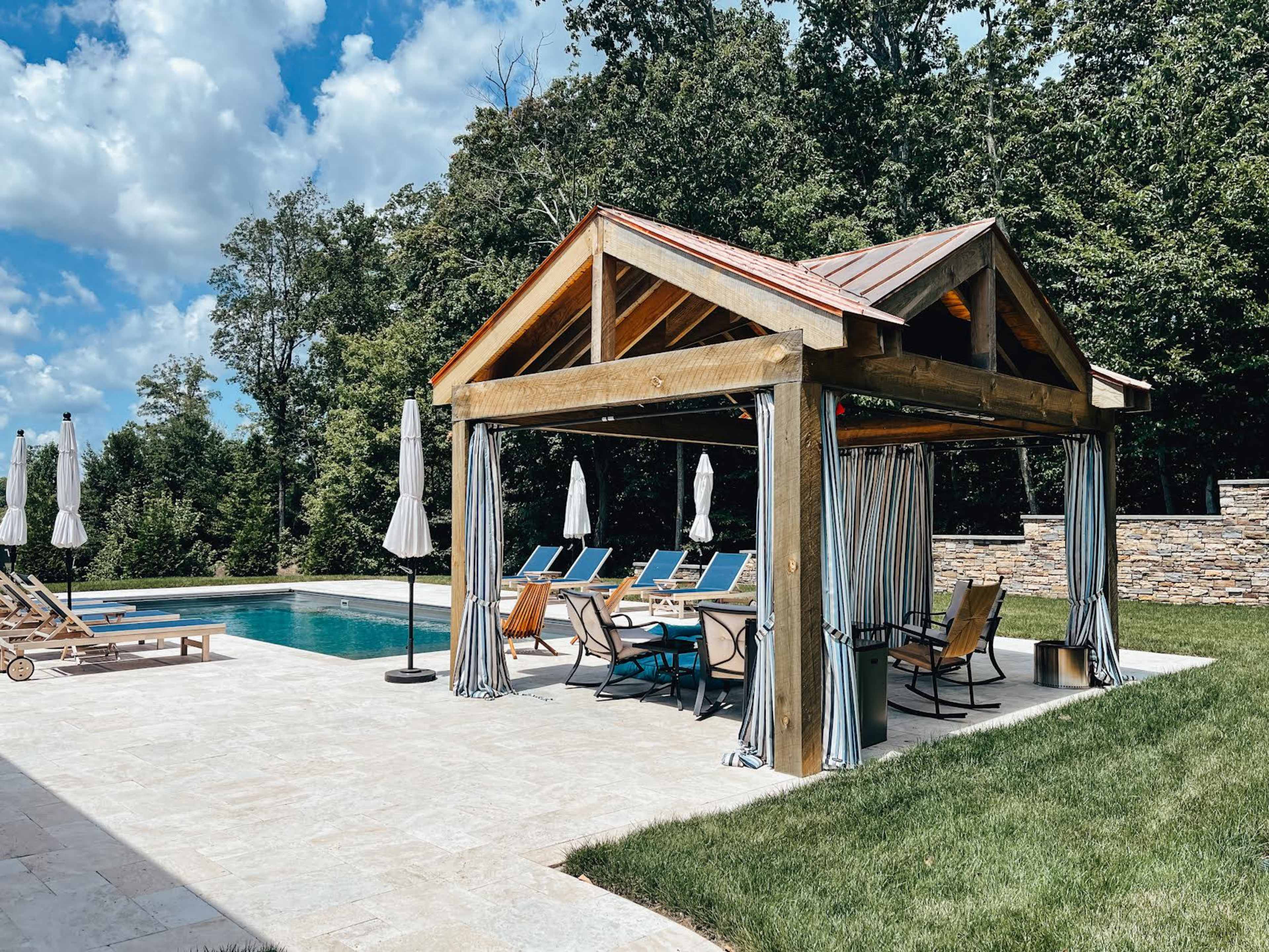 A wooden pavilion with a sloped roof and striped curtains shelters lounge chairs by a swimming pool surrounded by trees.