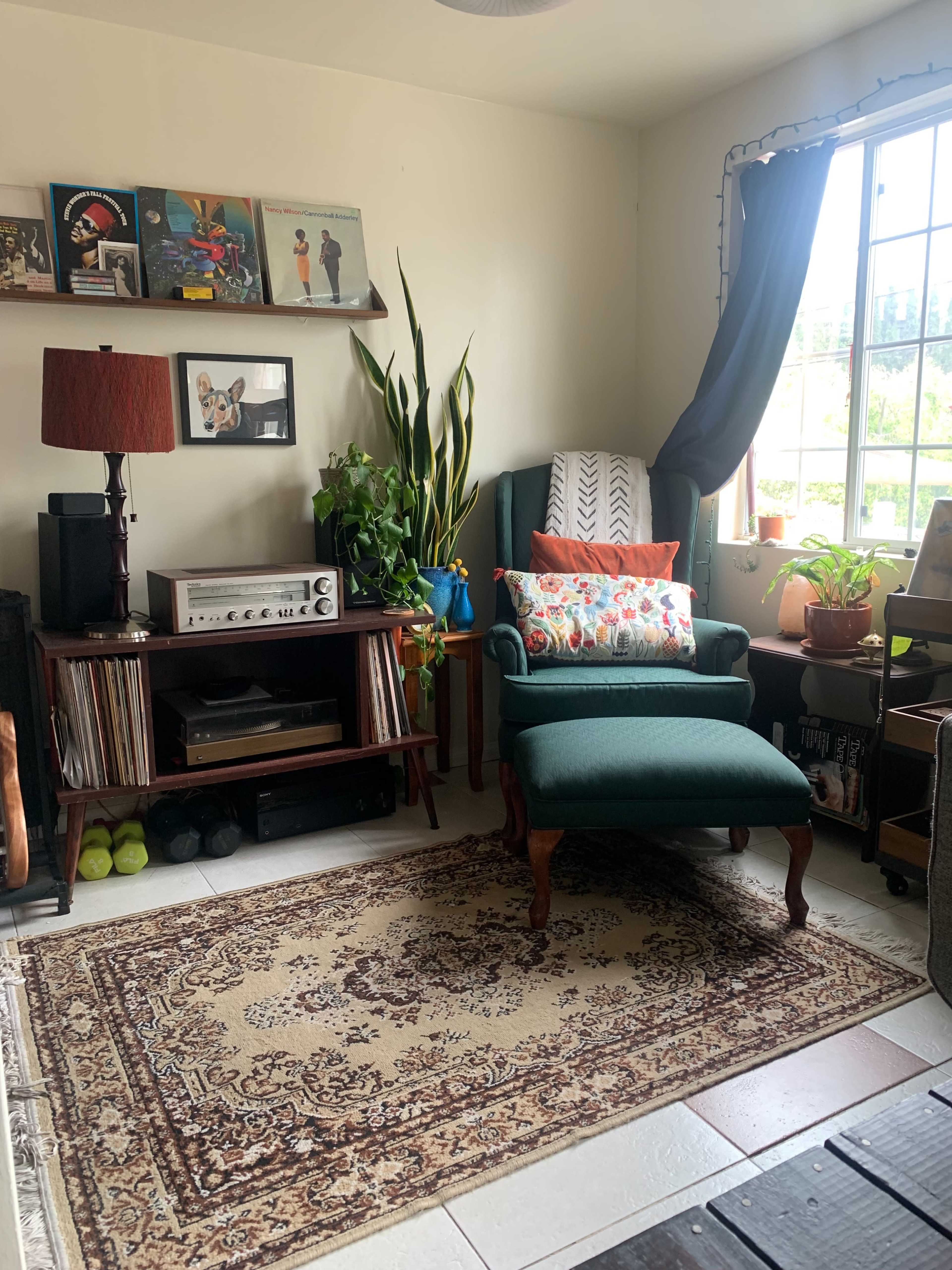 A cozy living space featuring a green armchair, a wooden shelf with vinyl records, plants, and a large window with curtains.