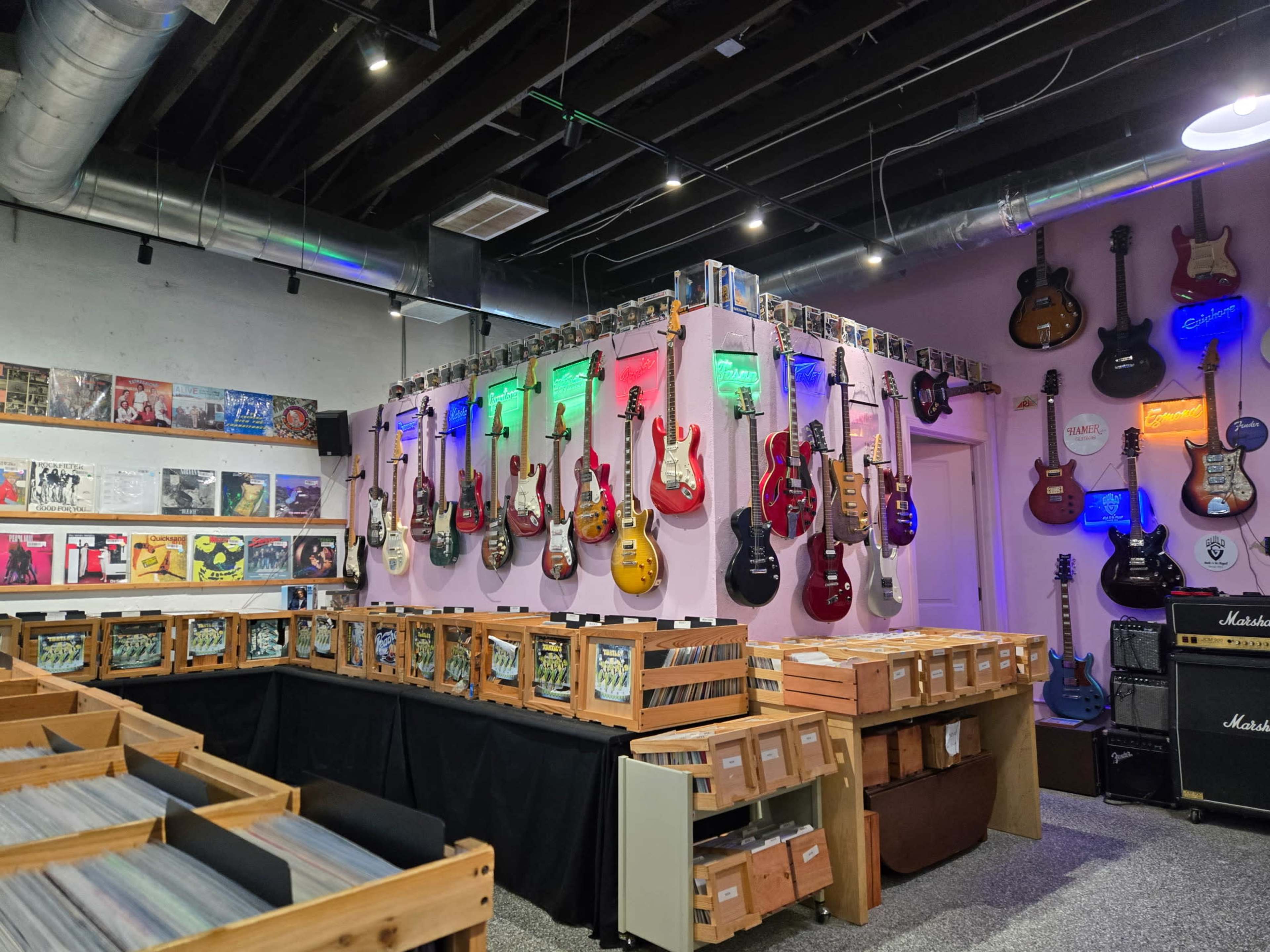 The image shows a music store featuring a wall of electric guitars illuminated with colorful lighting, surrounded by bins of vinyl records.