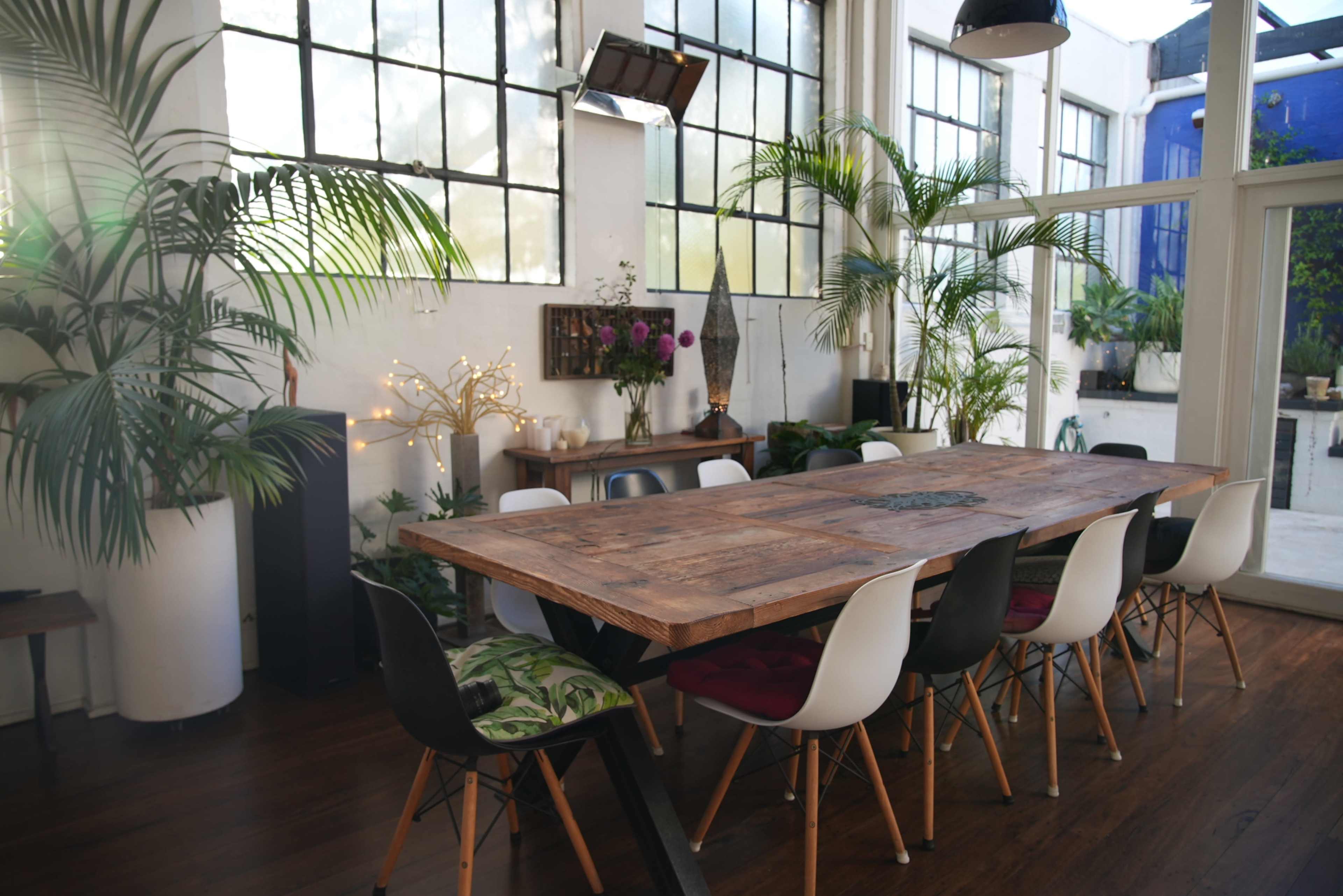 The image shows a large wooden dining table surrounded by various chairs in a bright, plant-filled room with large windows.