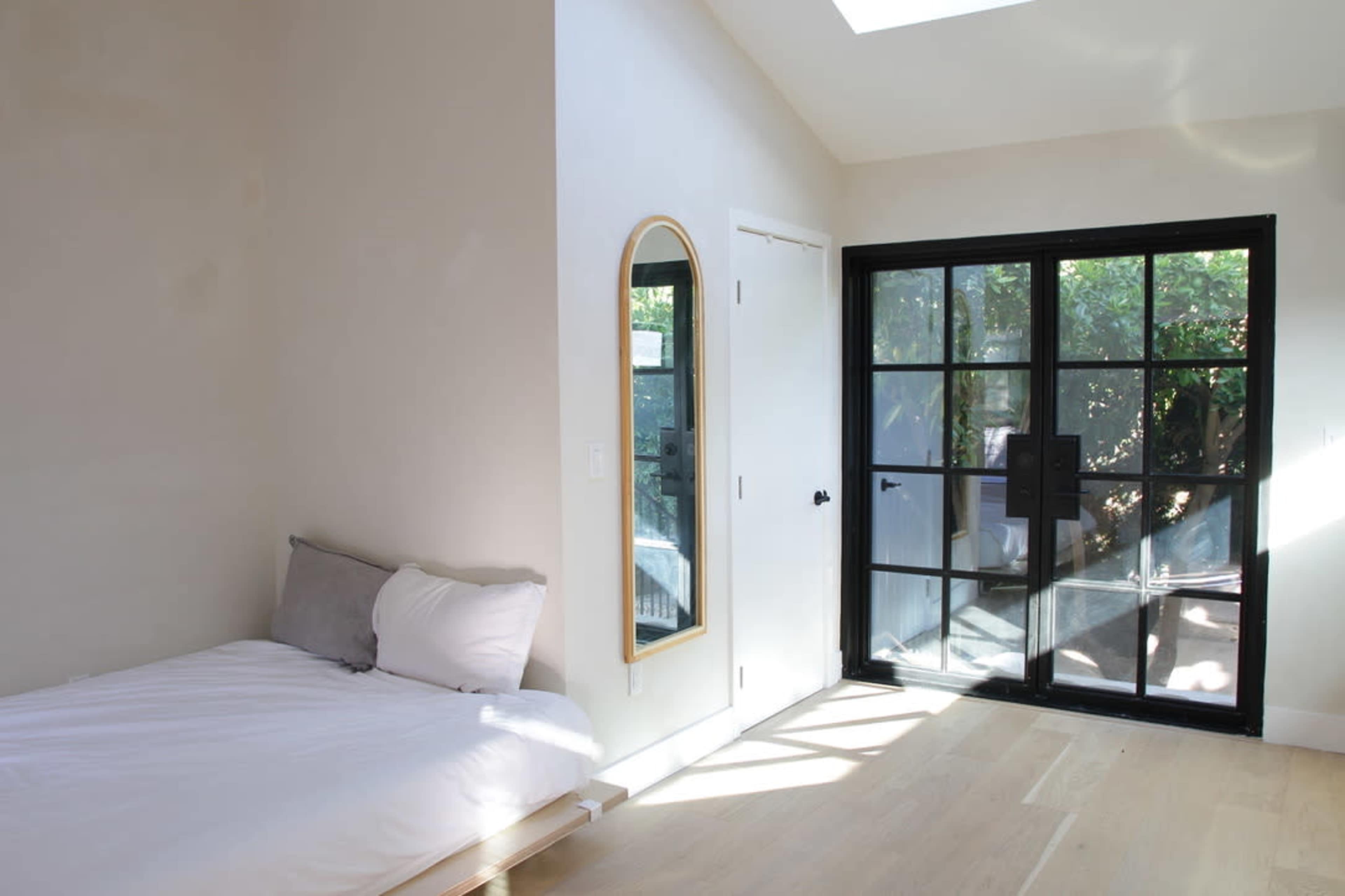 A small bedroom features a bed against one wall, a mirror, and a glass door leading to an outdoor area.