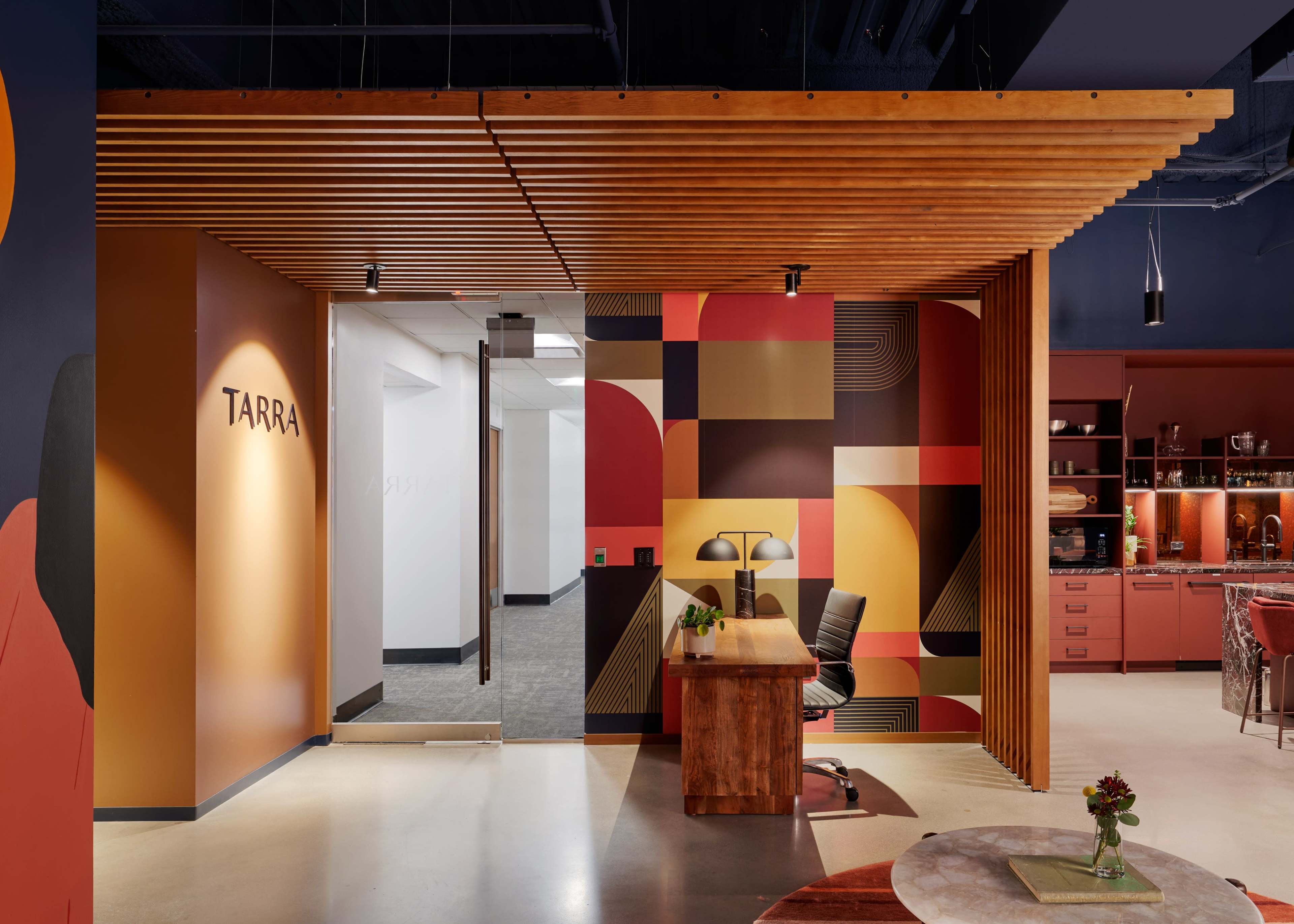 A modern office space with a wooden slatted ceiling, a desk with a black chair, and a colorful geometric wall design featuring shades of red, orange, and yellow.