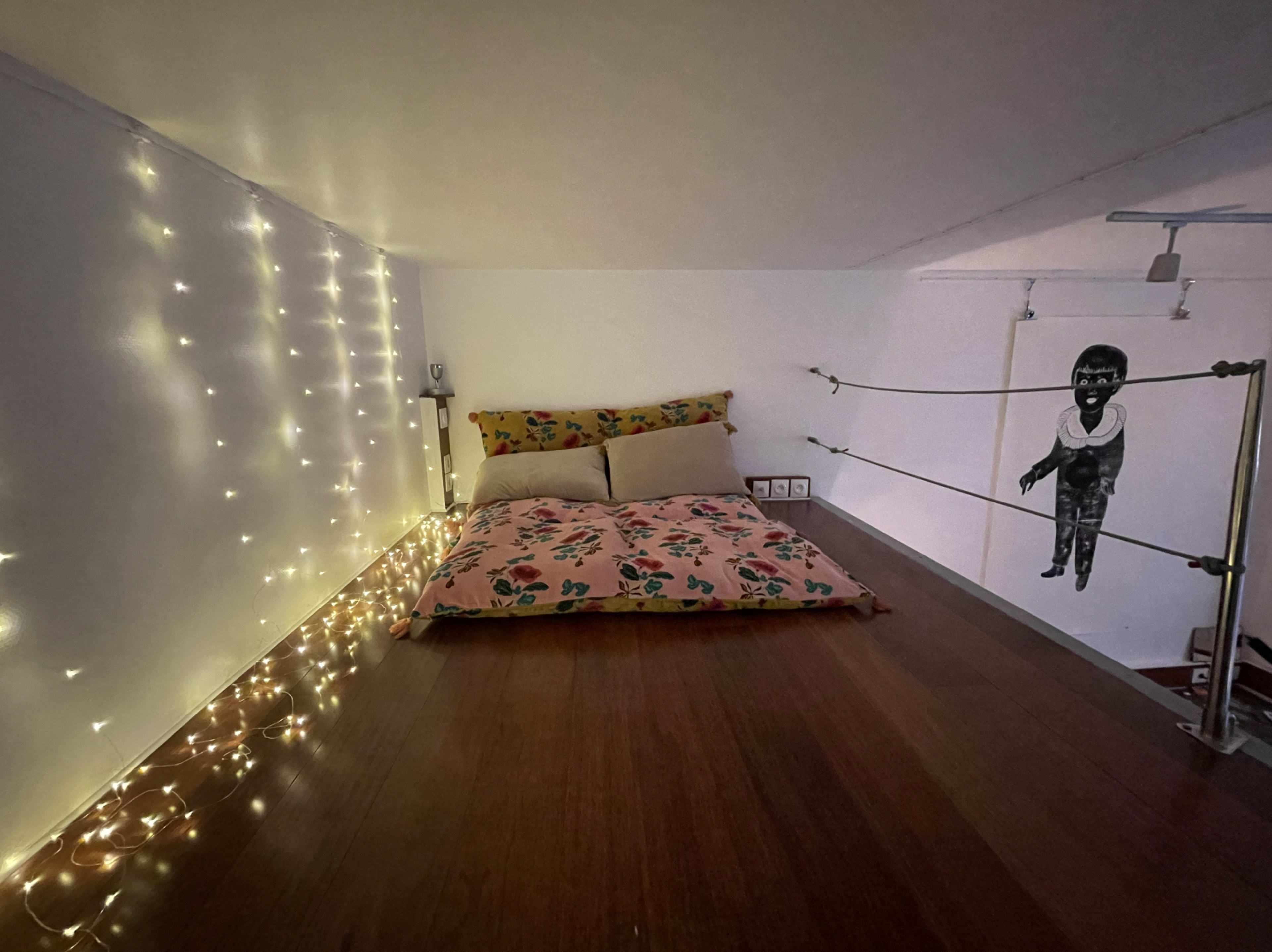 The image shows a small lofted sleeping area with a bed covered in floral-patterned bedding, string lights along one wall, and a black-and-white illustration of a child on the adjacent wall.