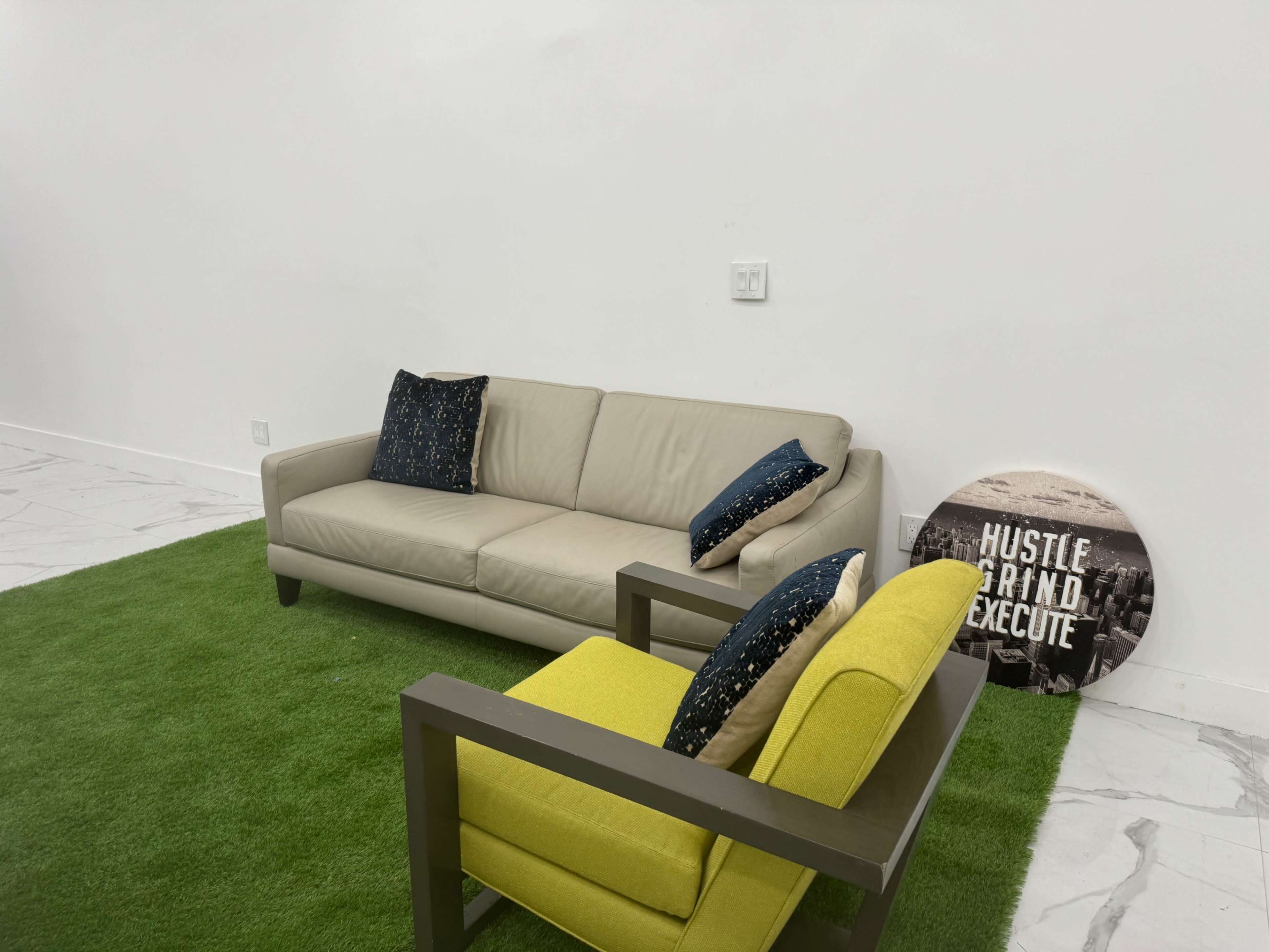 A beige sofa and a bright green chair are placed on a grassy rug in a minimalist room with white walls.