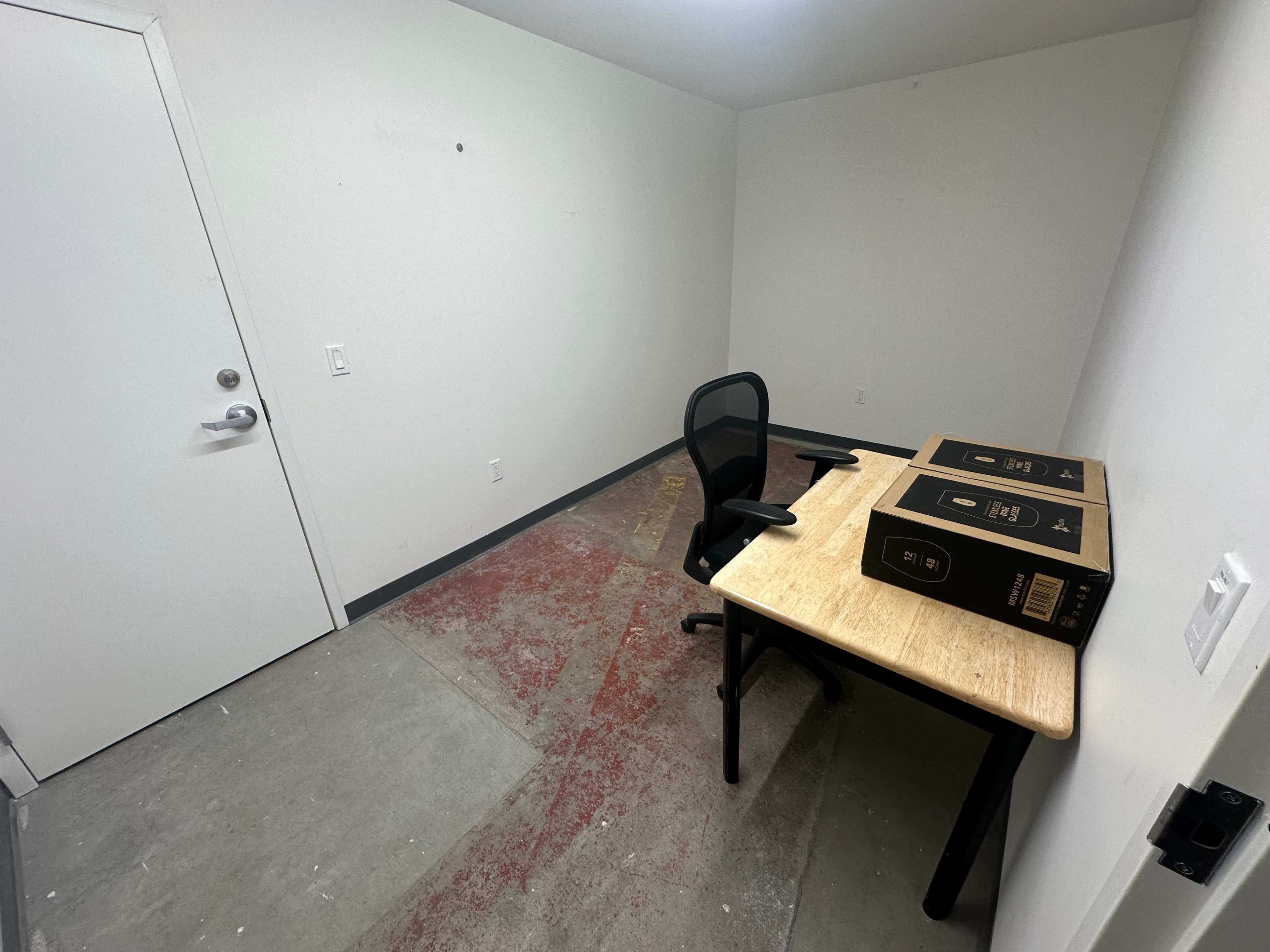 A small, empty room features a desk with two boxes on it and a chair positioned nearby.