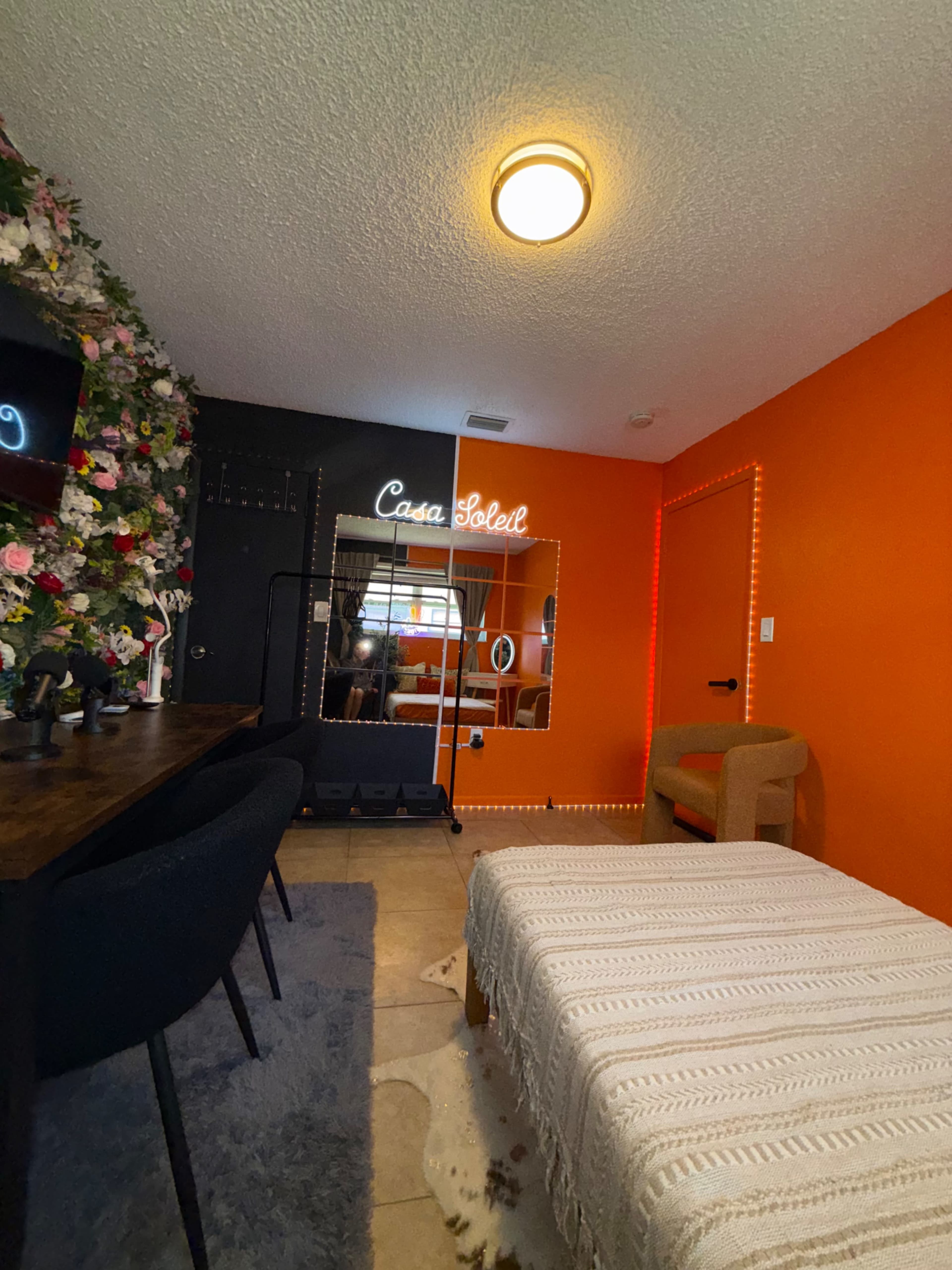The image shows a brightly colored room with an orange wall, a dressing table, a soft couch, and a mirror with neon signage that reads "Casa Soleil."