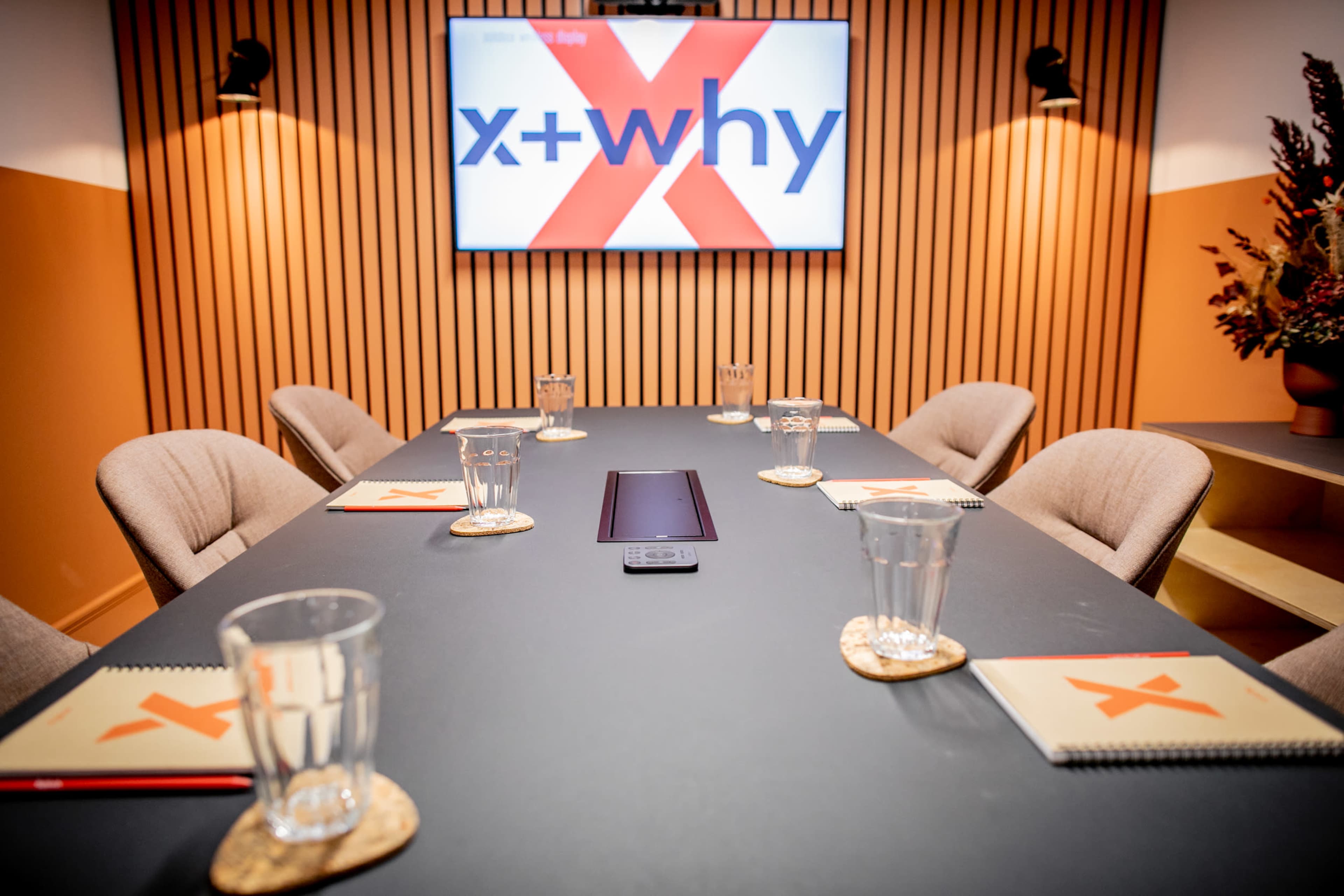 A modern conference room features a dark table set with notebooks and glasses, while a large screen displays a logo with the text "x+why."