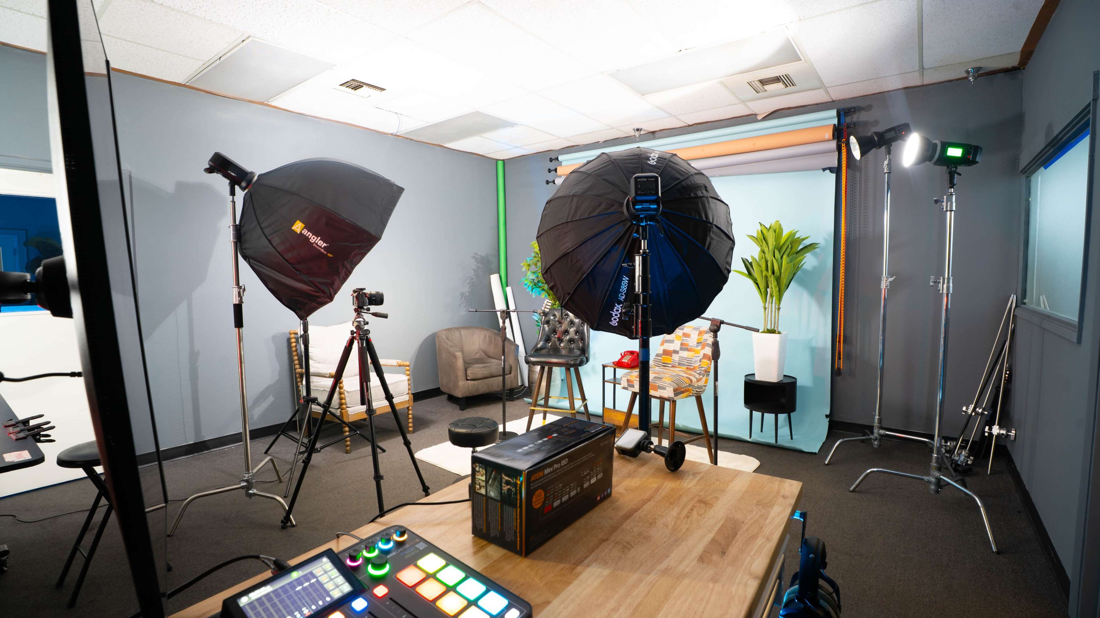 The image shows a photography studio setup with softbox lights, cameras on tripods, and a table adorned with various items in front of a backdrop.