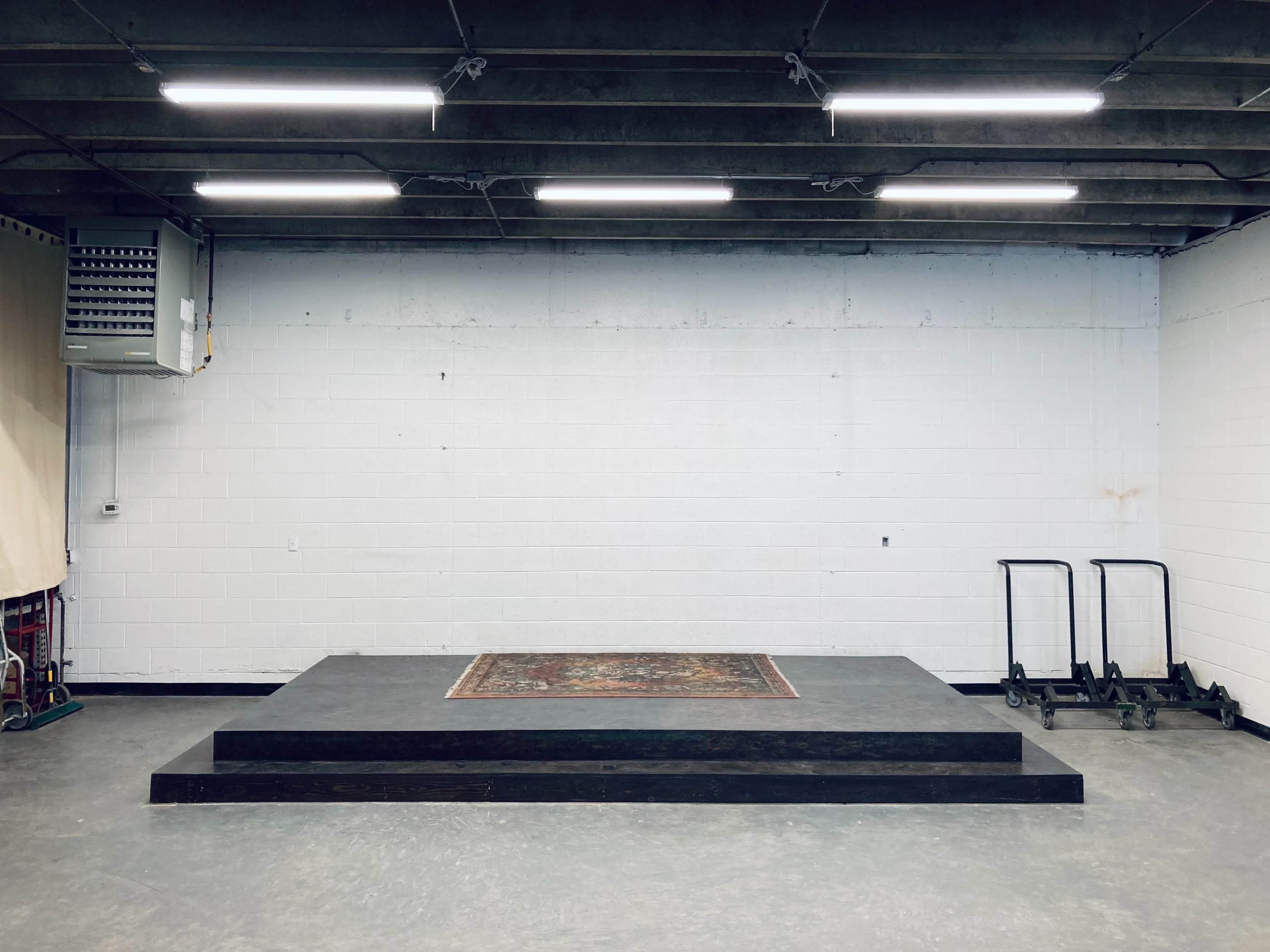 A blank stage with three steps and a decorative rug in the center, set against a plain white wall in a spacious room.