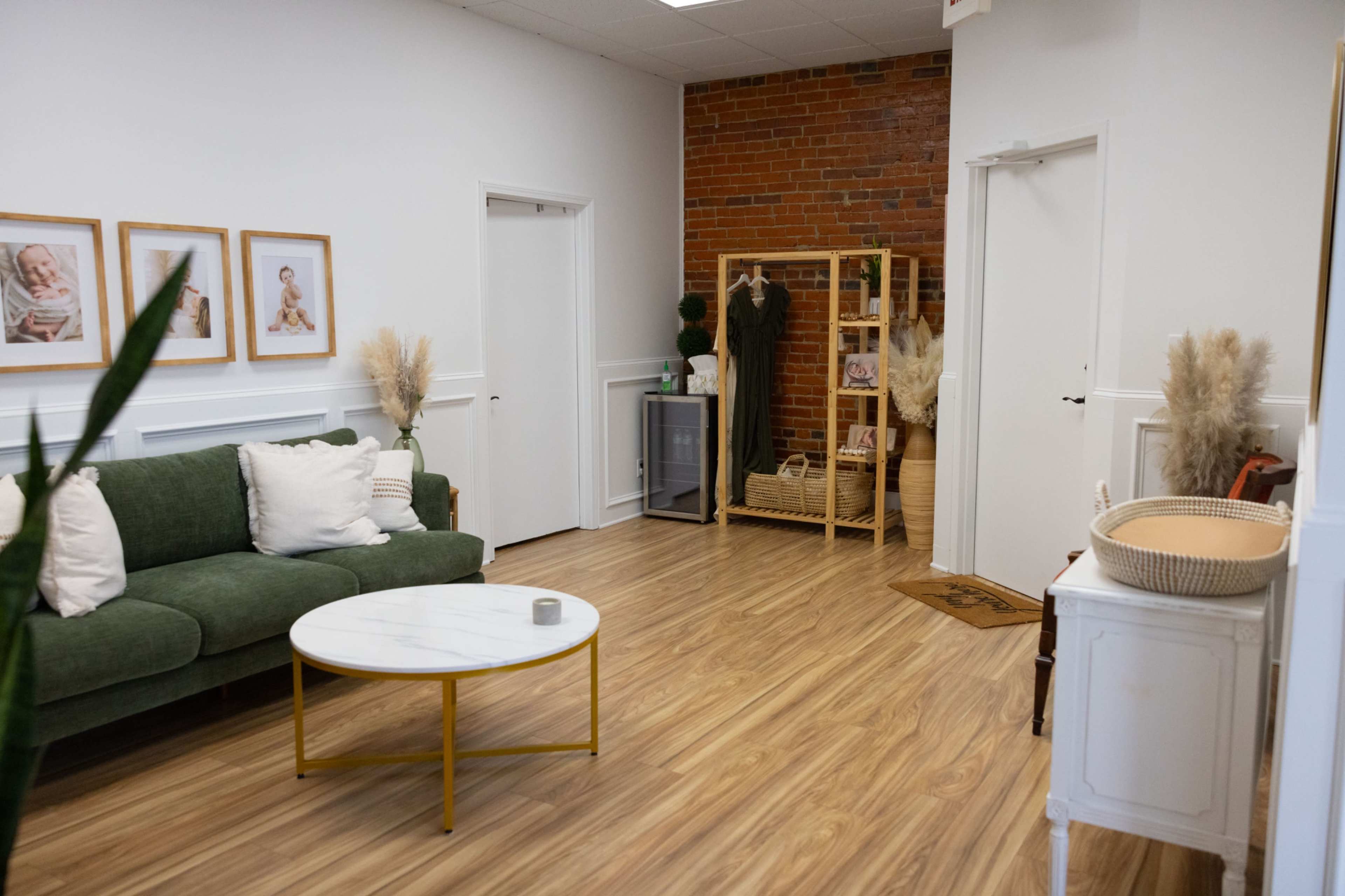 The interior of a room features a green sofa, a round marble coffee table, framed photographs on the wall, and a shelving unit against a brick wall.