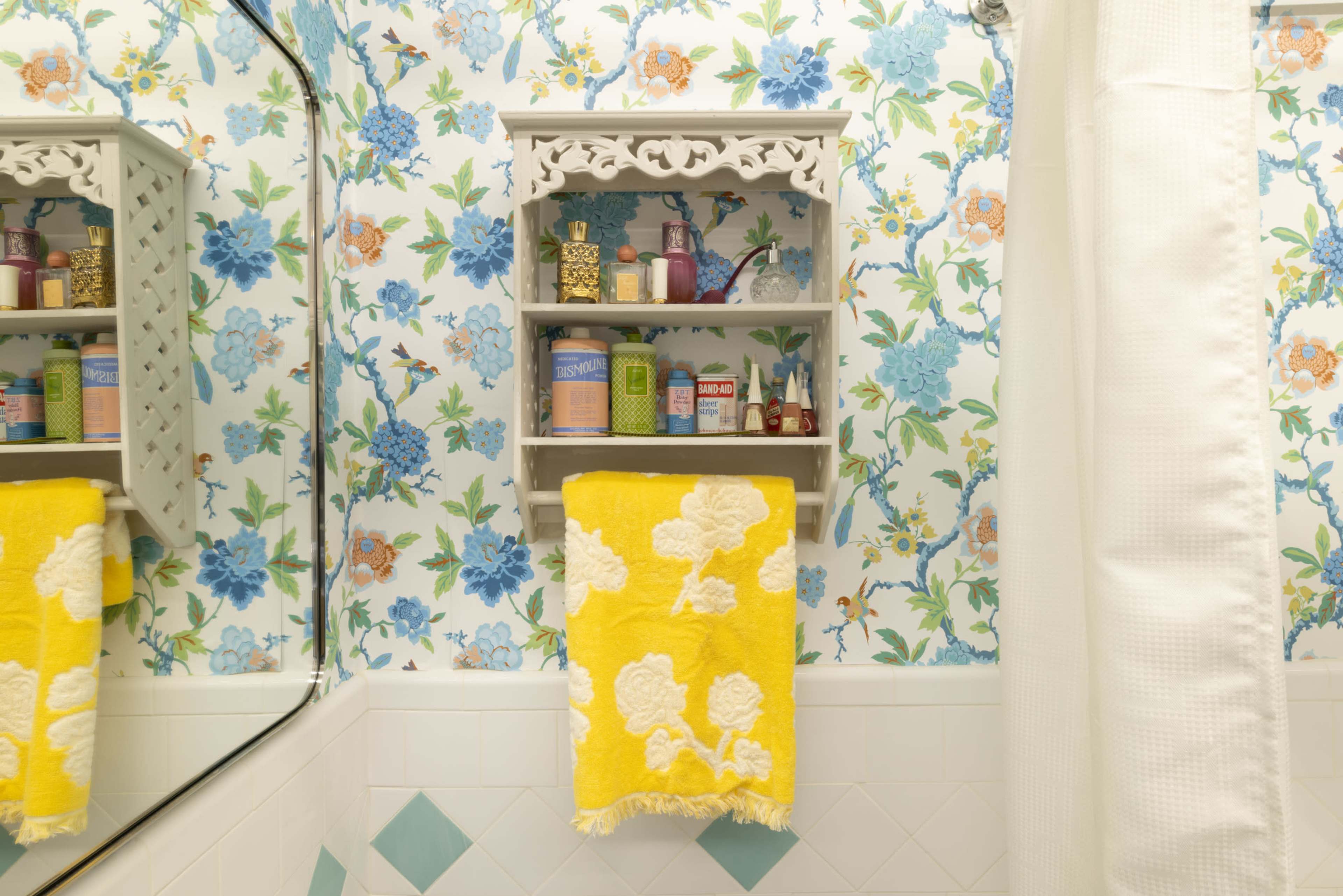A decorative bathroom shelf holds various toiletries against a floral wallpaper backdrop, with a yellow towel hanging below.