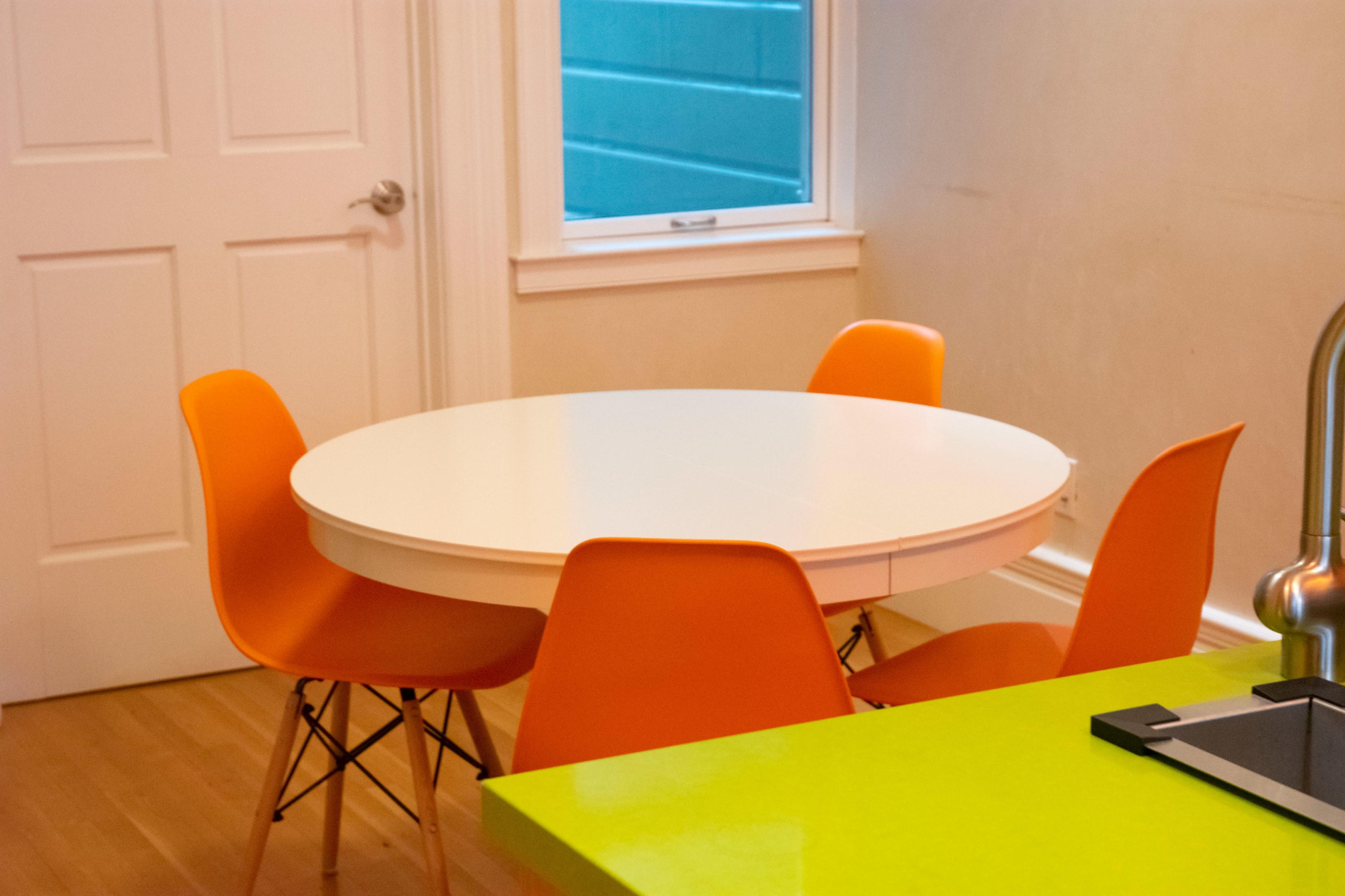 A round white table is surrounded by four orange chairs in a brightly lit room with a window and a closed door.