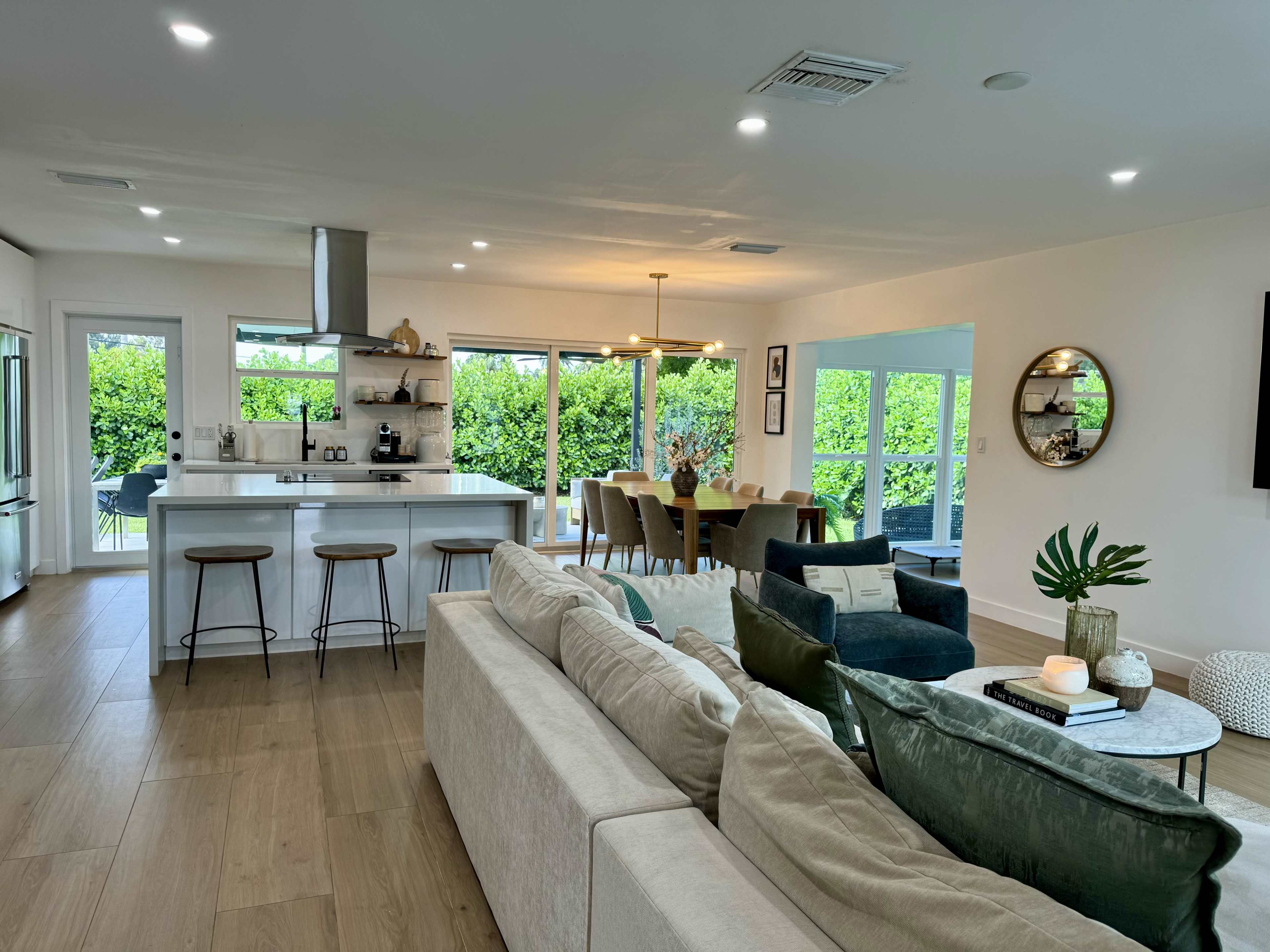 The image shows a modern open-concept living area featuring a beige sofa, a dining table with chairs, and a kitchen with a large island, surrounded by large windows overlooking greenery.