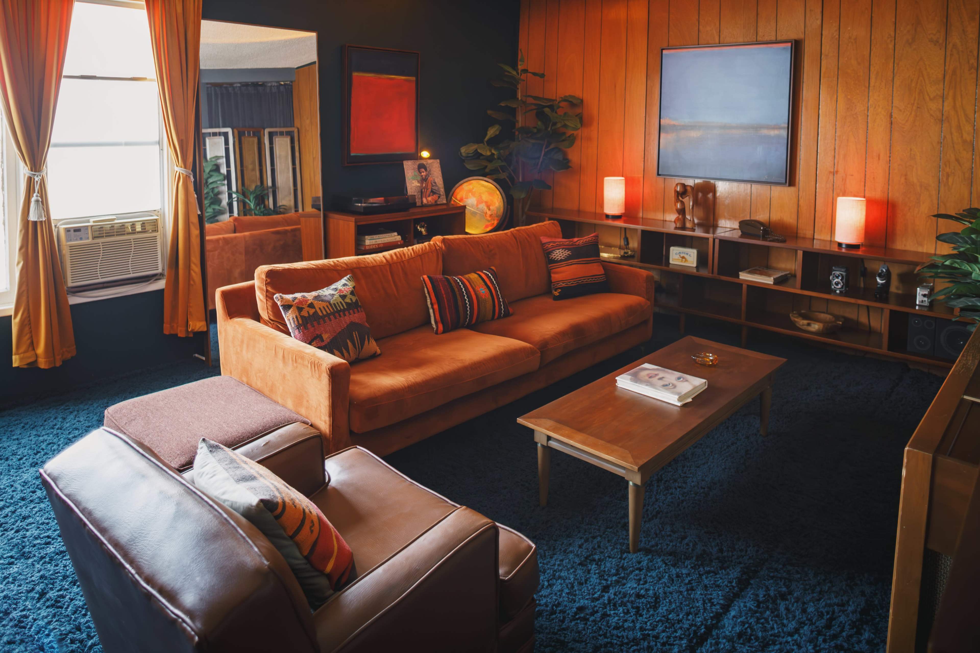 A retro-style living room featuring an orange sofa, a wooden coffee table, and plants, with a combination of warm lighting and dark walls.