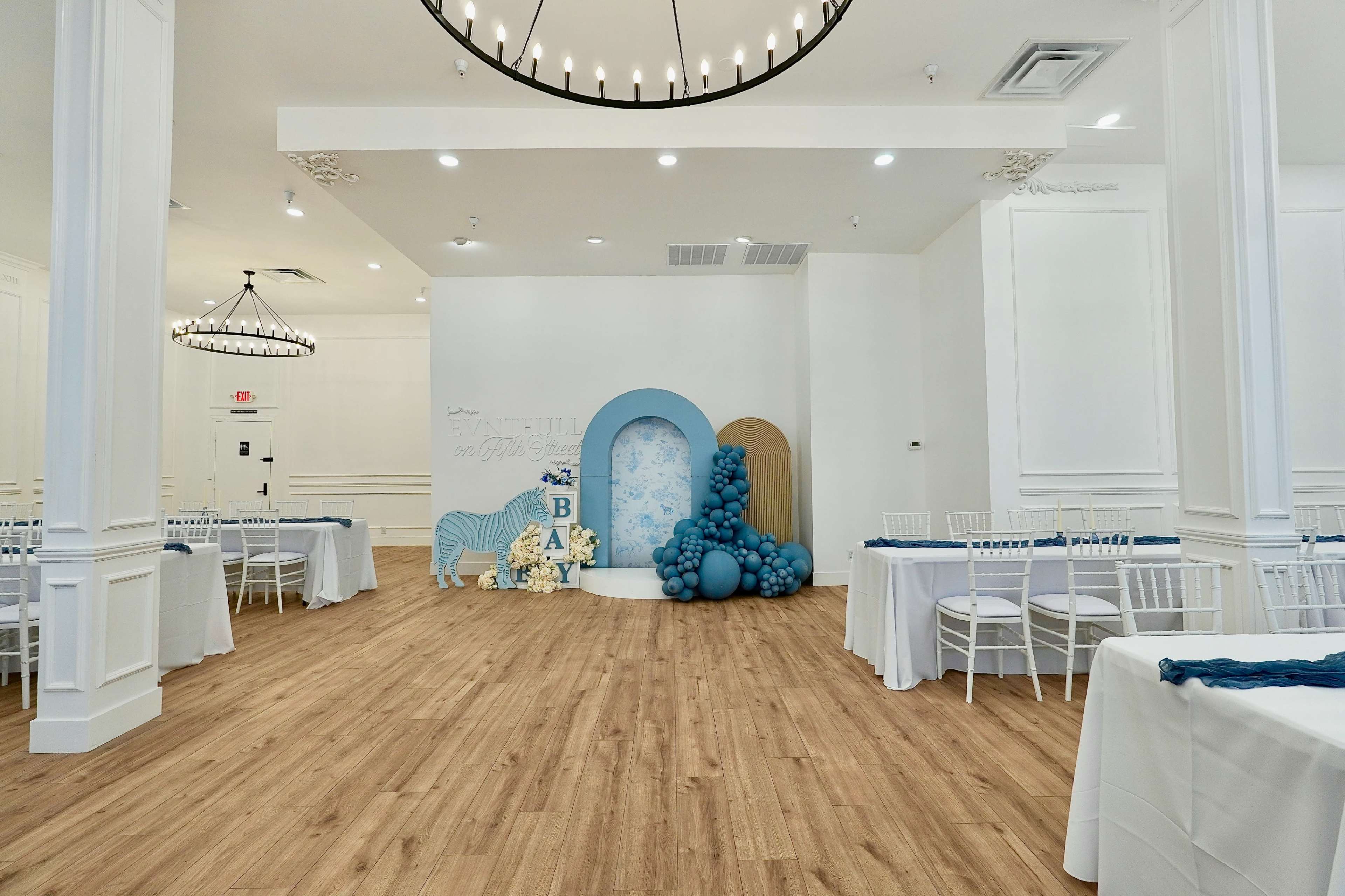 The image shows a spacious event room with white walls, wooden flooring, and tables set with blue tablecloths, featuring decorative balloons and a backdrop for an event.