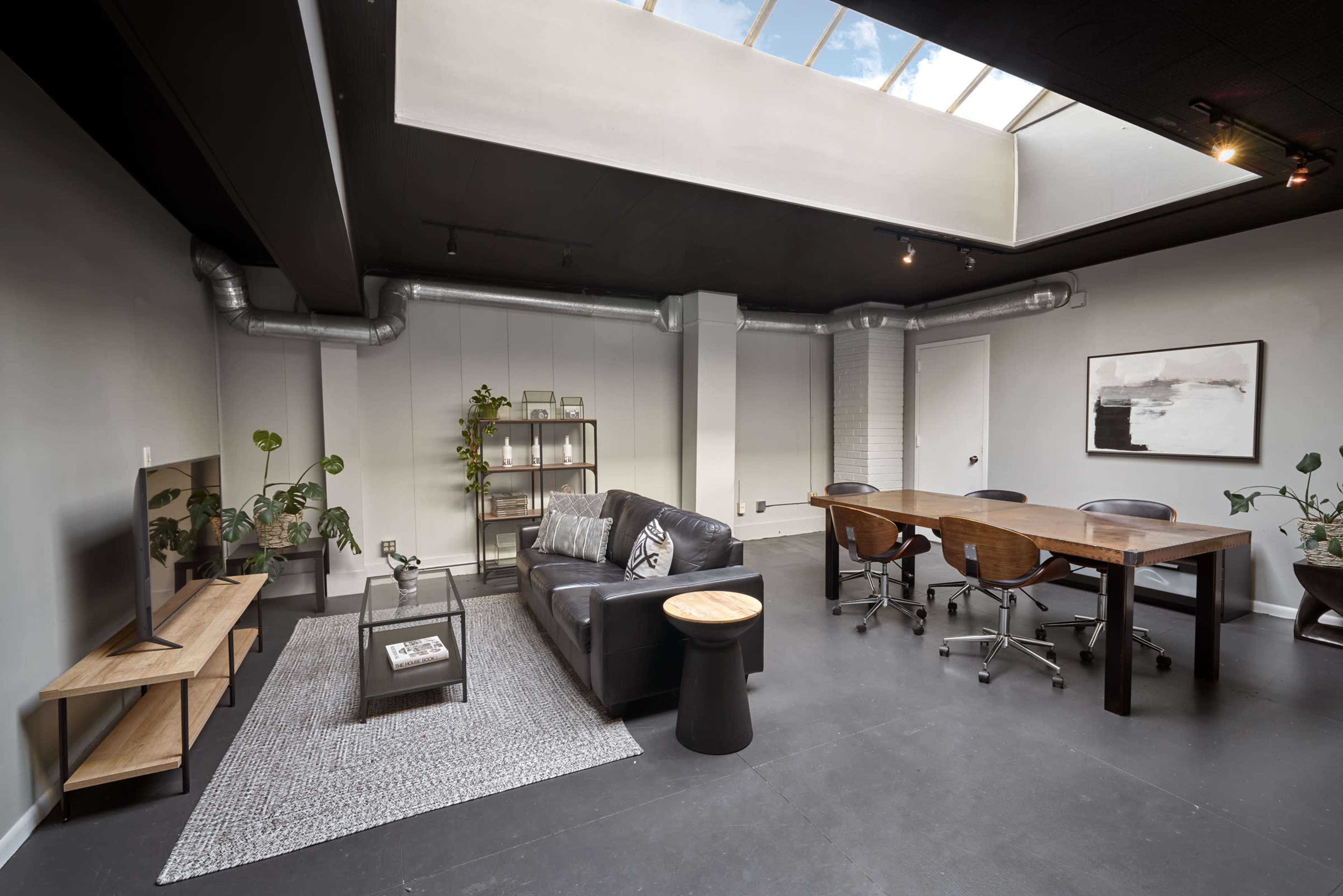 The image shows a modern office space featuring a black leather sofa, a wooden dining table with chairs, plants, and a skylight that brightens the room.