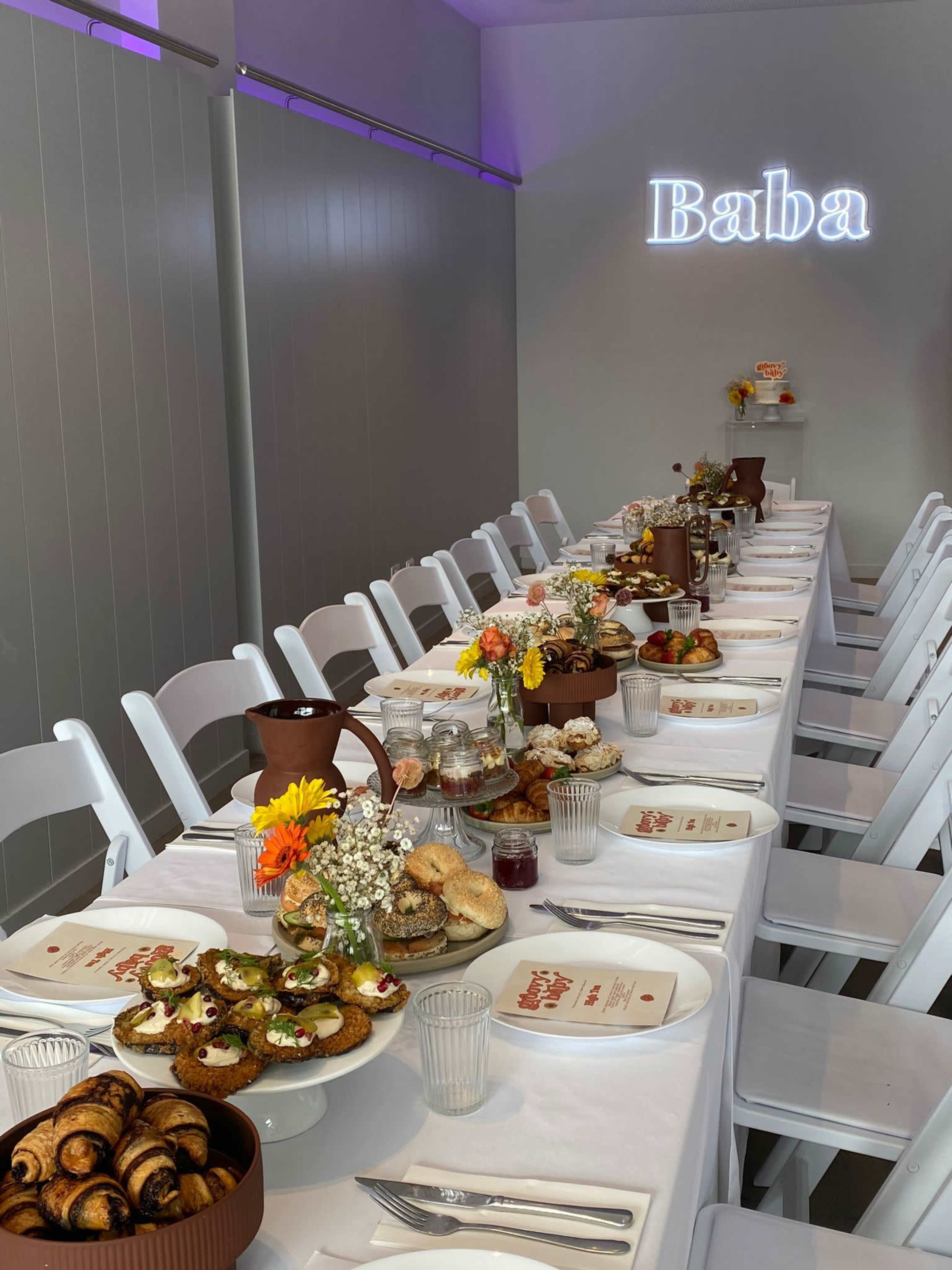 A long dining table is set with various dishes and decorated with flowers, under a neon sign that reads "Baba."