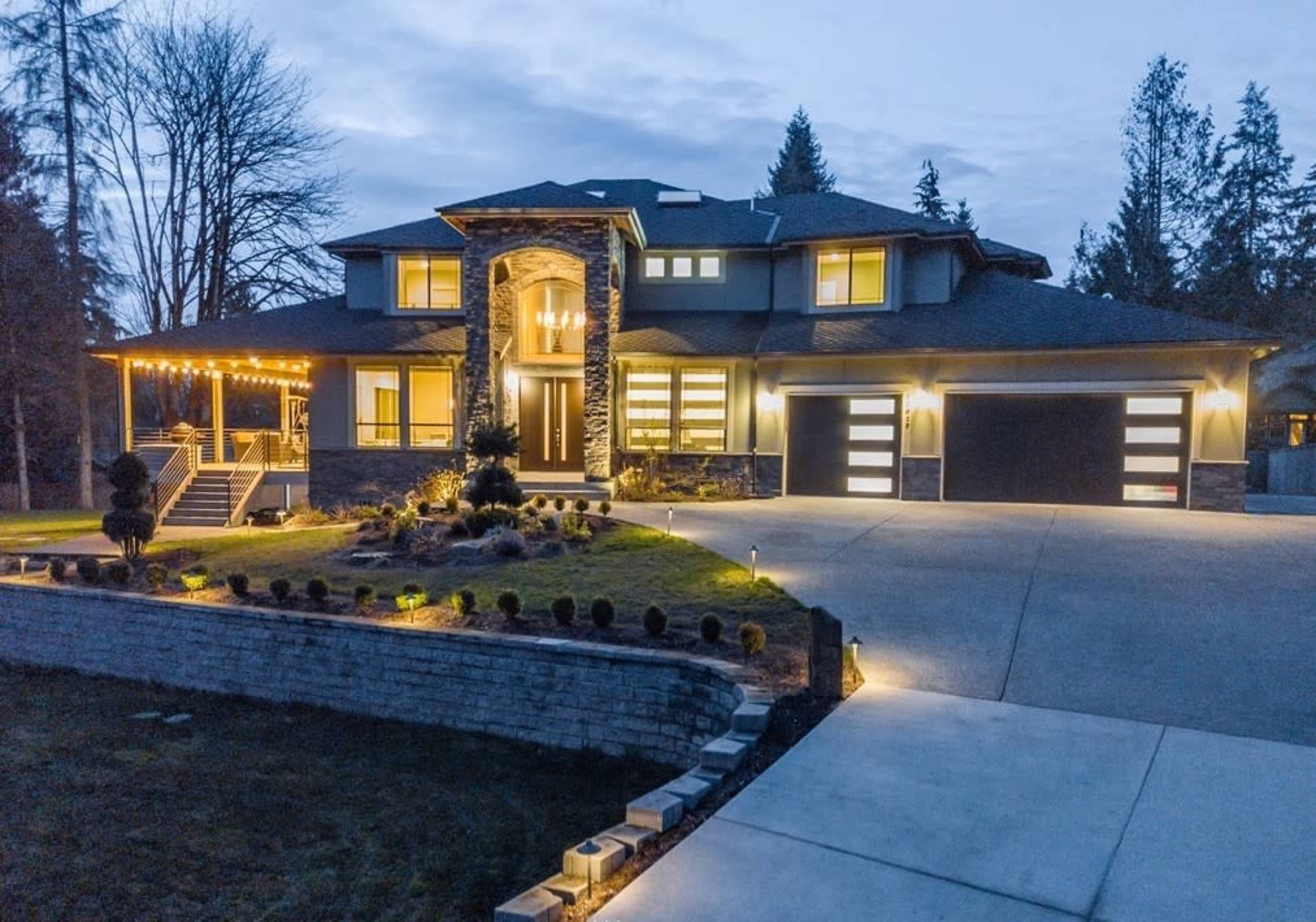 A modern two-story house with stone accents and exterior lighting, featuring a landscaped yard and a driveway leading to three garage doors.