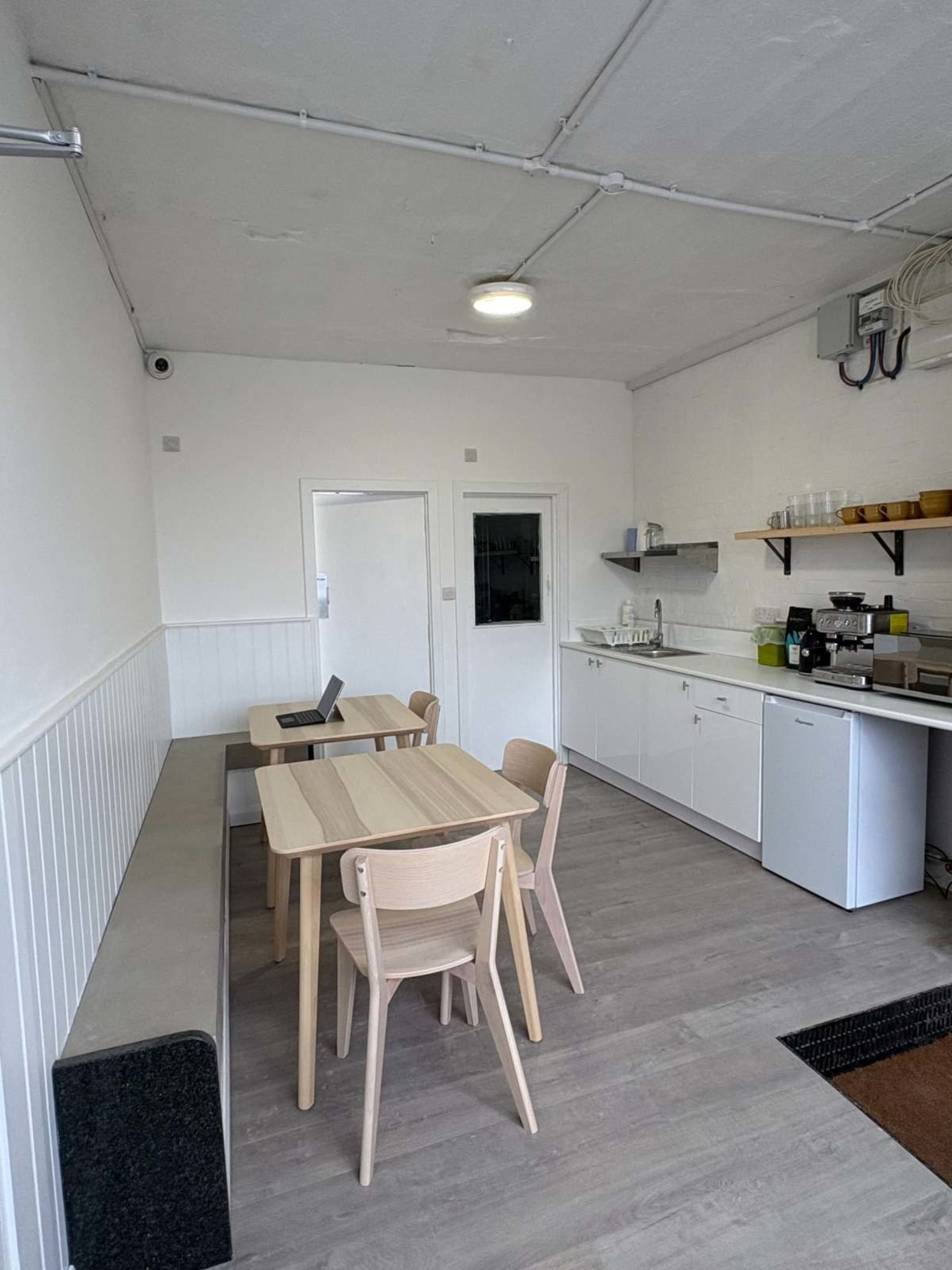 The image shows a small, modern kitchen area with a dining table, chairs, and appliances along the walls.