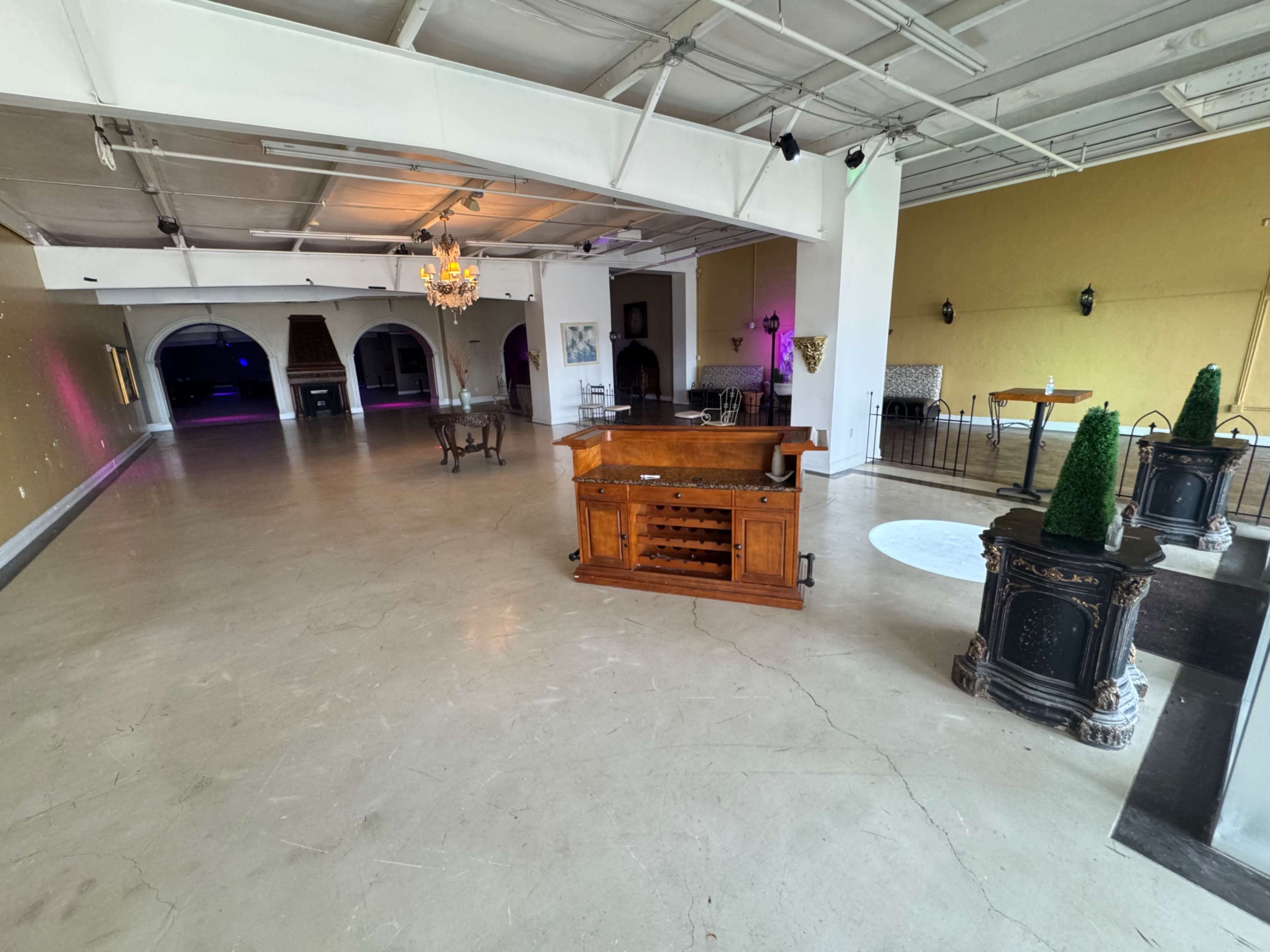 The image shows a spacious, empty interior with hardwood and concrete flooring, featuring a wooden bar in the center and various seating areas around the room.