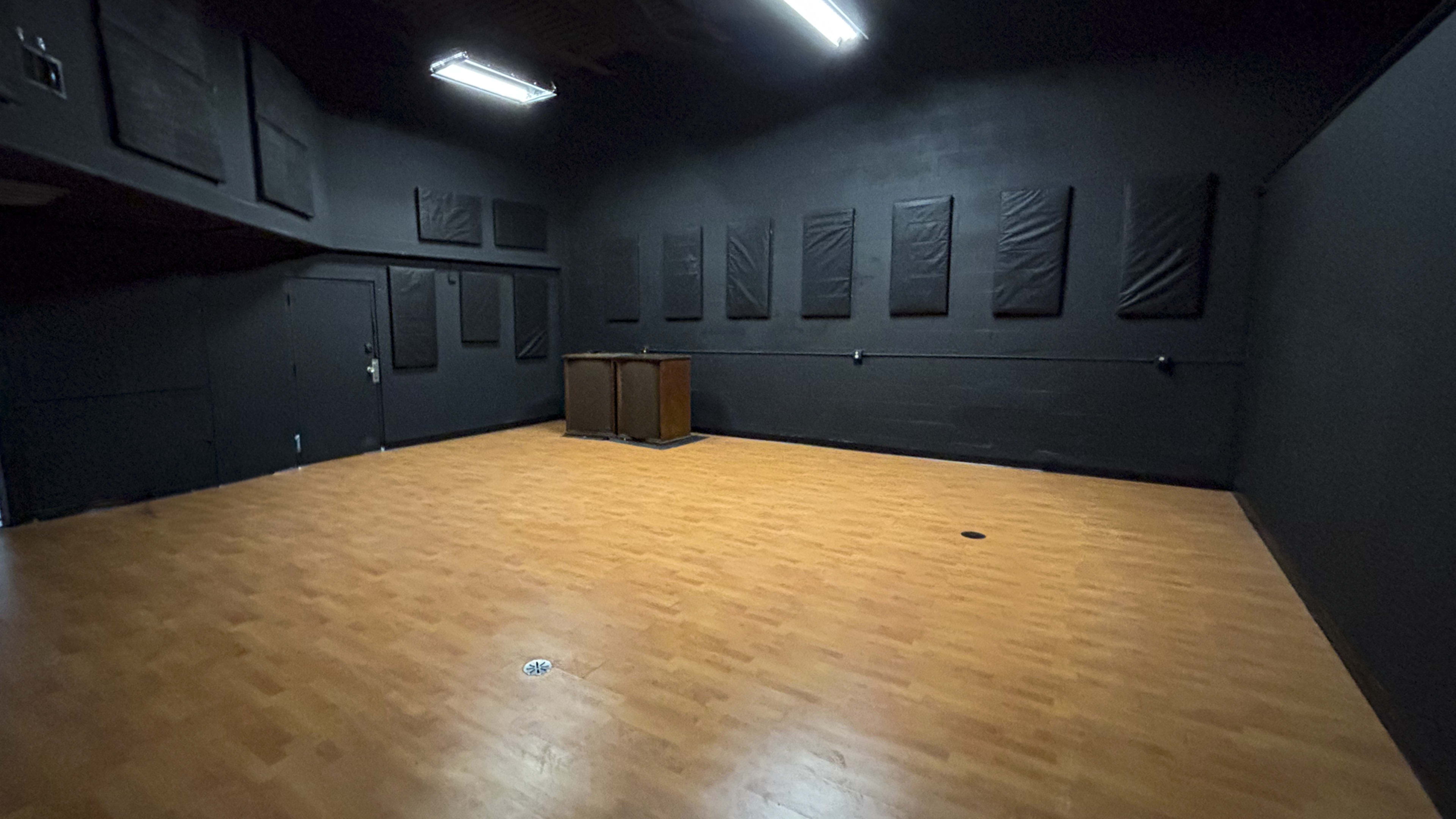 The image shows a large, empty room with black walls, wooden flooring, and a couple of tables at the center.