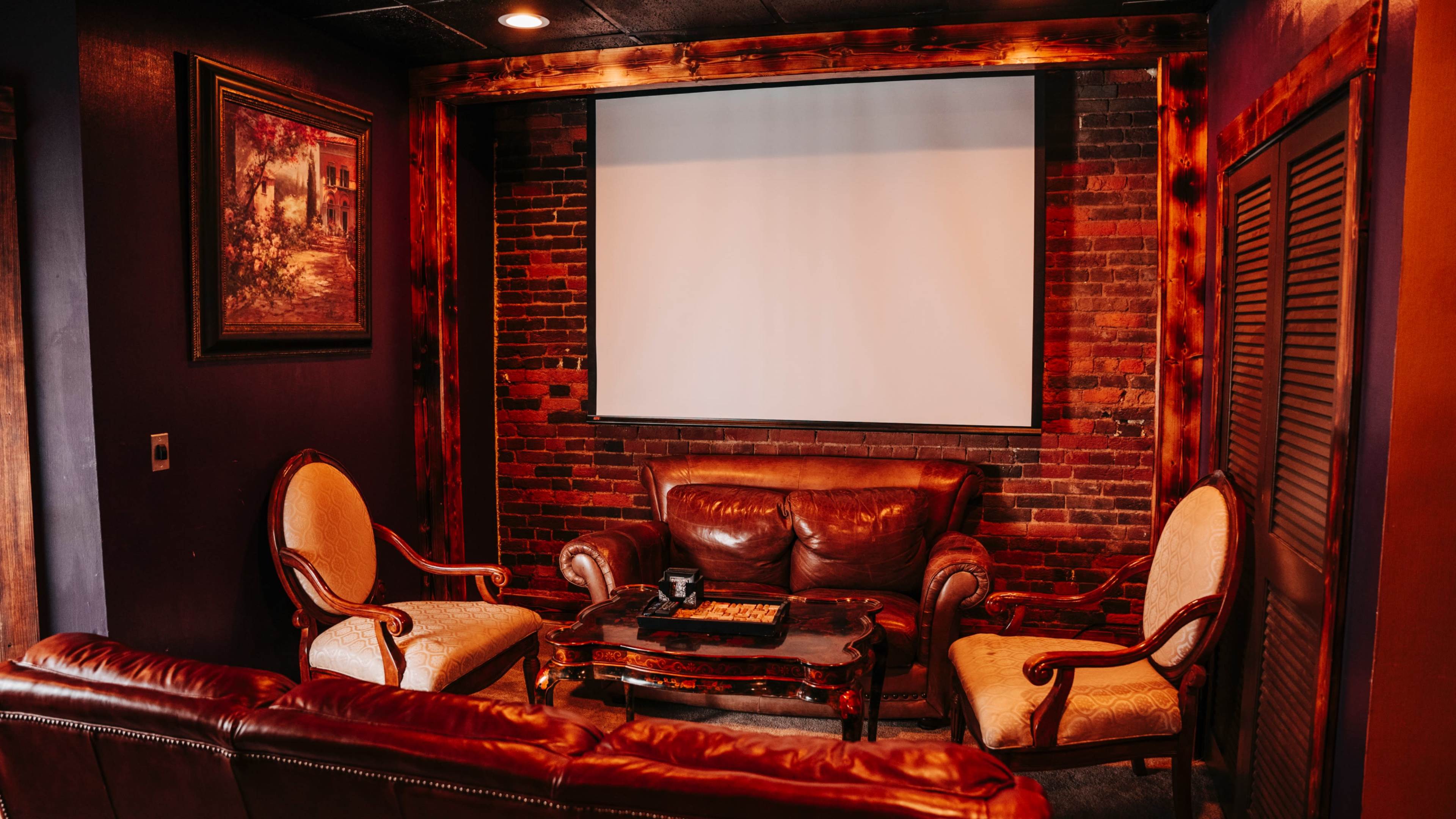The image shows a cozy, dimly lit room with a leather sofa and armchairs arranged around a coffee table, facing a blank projection screen and exposed brick walls.